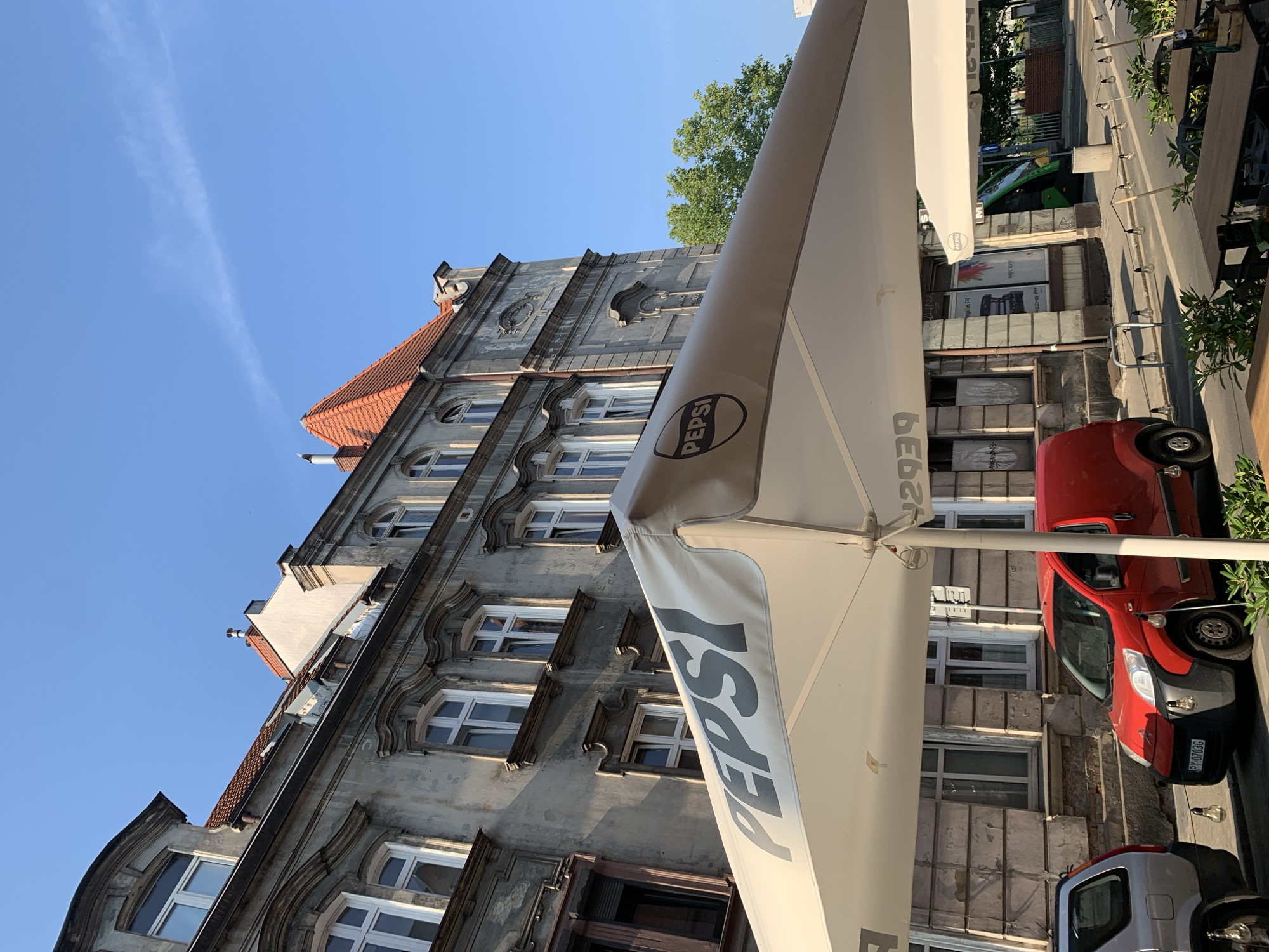 This is a street-level, outdoor photograph taken in the city of Poznan, Poland. The scene is dominated by the facade of an old, weathered building with ornate architectural details and multiple windows. In the foreground, a large white umbrella with the "PEPSI" logo is prominently displayed, casting a shadow. A red van is parked on the street in front of the building, and a silver SUV is partially visible to the left. The street is paved, and there are tables and chairs set up under the umbrella, suggesting an outdoor seating area for a cafe or restaurant. The sky is clear and blue with a faint contrail. The overall impression is of a sunny day in an urban environment with historical architecture.