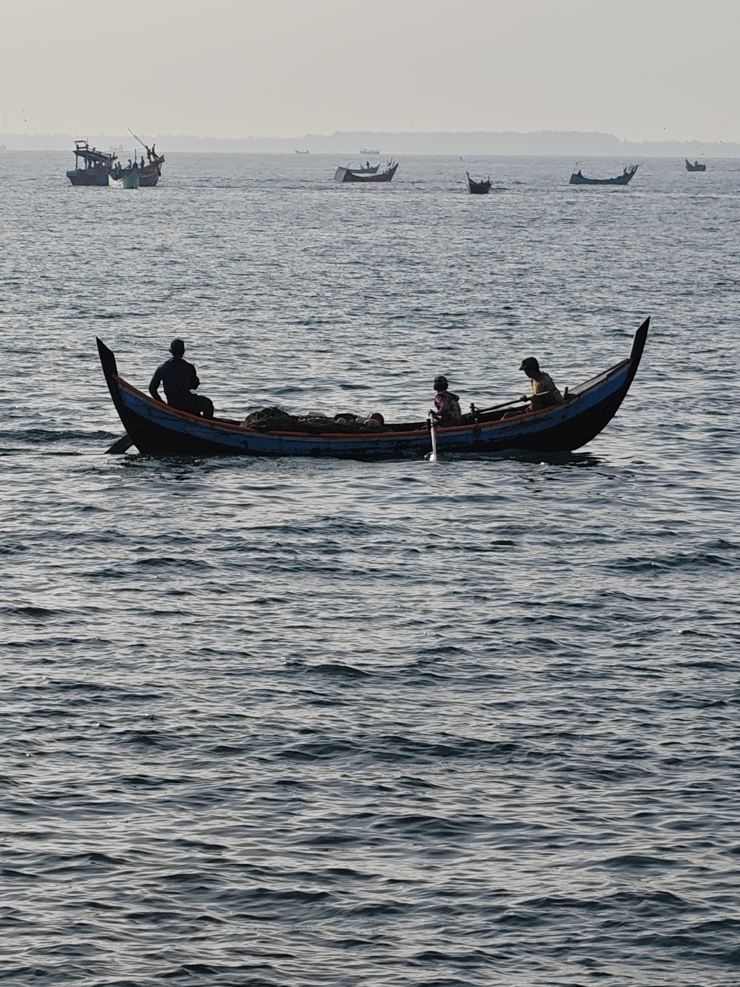 This photograph depicts a tranquil maritime scene featuring several traditional boats on the water in Lhokseumawe, Indonesia.
In the foreground, a wooden boat, characterized by its distinct pointed bow and stern and painted with blue and orange accents, is occupied by three people. The individuals are silhouetted against the brighter sky, making their features indistinct. One person sits at the stern, another in the middle, and a third at the bow, holding an oar. Fishing nets and other equipment are visible stowed within the boat. The boat appears to be moving gently across the water.
In the mid-ground and background, numerous other traditional fishing boats are scattered across the sea, also appearing as silhouettes. Some are smaller, similar in style to the foreground vessel, while others are larger, more substantial fishing vessels with structures on their decks. The water is calm with gentle ripples reflecting the soft daylight. The horizon is a light, hazy band, suggesting either early morning or late afternoon, and a faint landmass can be seen in the far distance. The overall atmosphere is peaceful and serene, illustrating daily life and work on the water. No text is visible in the image.