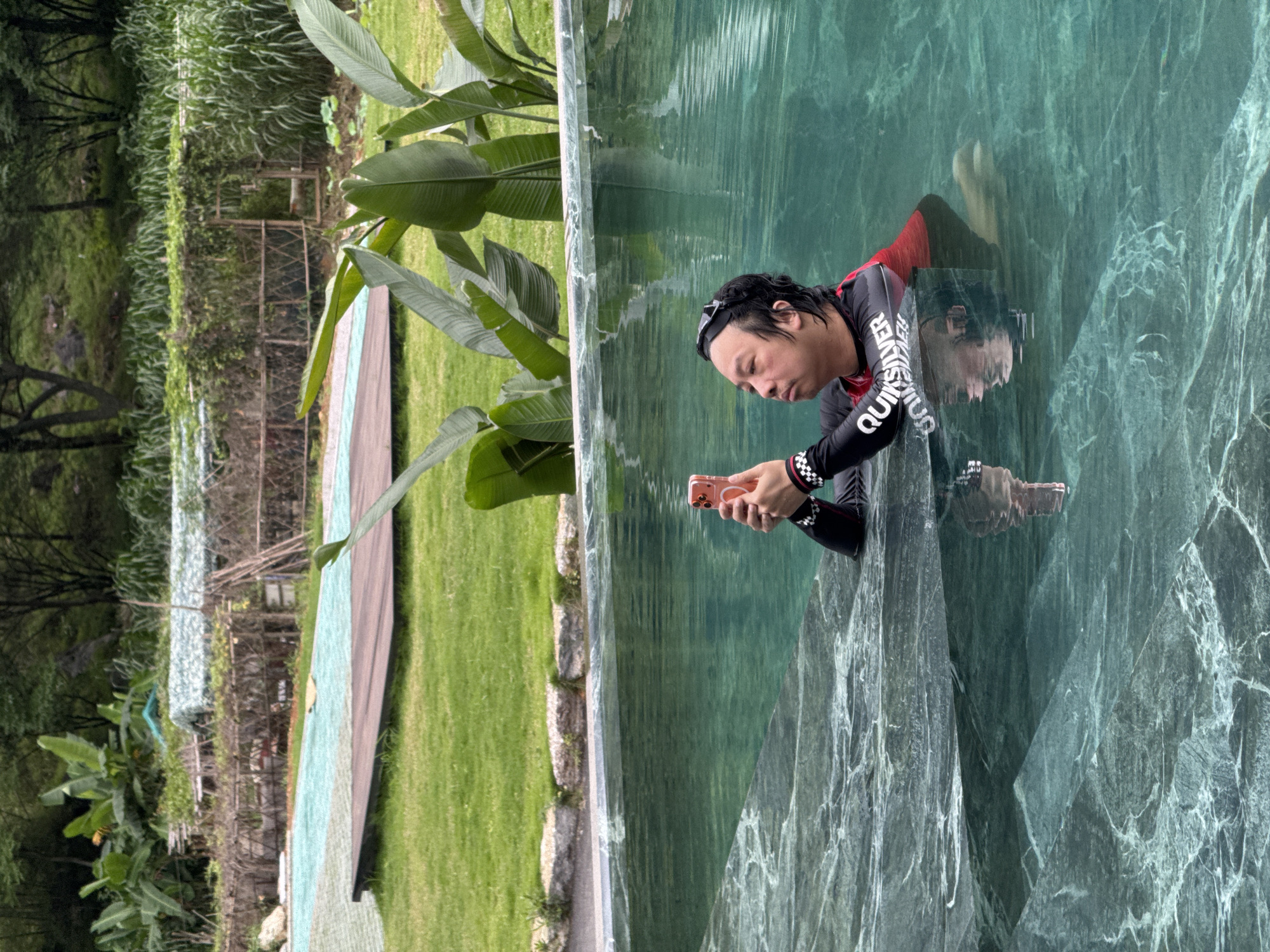 A man in a black and red long-sleeved wetsuit with "Quiksilver" printed on the arm is looking at a pink smartphone while partially submerged in a green-tinted pool. The pool's edge is made of a green marble-like material. Reflected in the water is the man and the phone. Behind the pool, there is a lush green lawn and tropical plants. Further back, there is a wooden fence and trees. The location is Guilin, China.