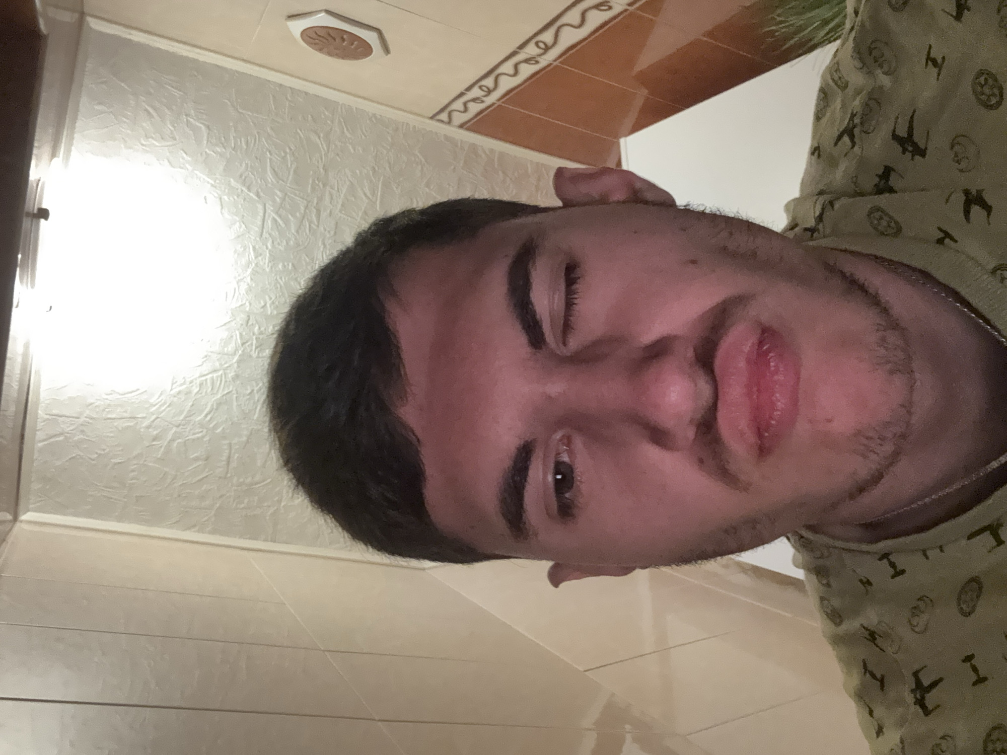 This is a vertically oriented selfie of a young man, likely in his late teens or early twenties, taken indoors in Burgas, Bulgaria. The man has short, dark hair and prominent eyebrows. He has dark eyes and a light amount of facial hair (stubble) on his chin and upper lip. His lips are slightly parted, giving him a somewhat pouting or neutral expression, and he is looking directly at the camera.
He is wearing a light olive green or beige t-shirt adorned with a pattern of Star Wars-themed symbols, including X-wing fighters, TIE fighters, and circular emblems resembling the Galactic Empire or Rebel Alliance logos. A thin silver-colored chain is visible around his neck.
The background reveals interior details of what appears to be a room, possibly a bathroom or hallway. The wall behind him has a light color and a distinct, textured surface, possibly stucco or embossed wallpaper. Above this, there's a strip of brownish-orange rectangular tiles with a wavy, darker brown decorative border. Higher up, on what seems to be the ceiling or a soffit, there's a light-colored hexagonal or octagonal fixture with a spiral or sun-like pattern in the center. Below the textured wall, lighter, possibly beige, horizontal tiles are visible. A bright, artificial light source illuminates the left side of the frame, creating a strong glare on the textured wall. Some blurred green foliage is visible in the upper right background. No visible text is present in the image.