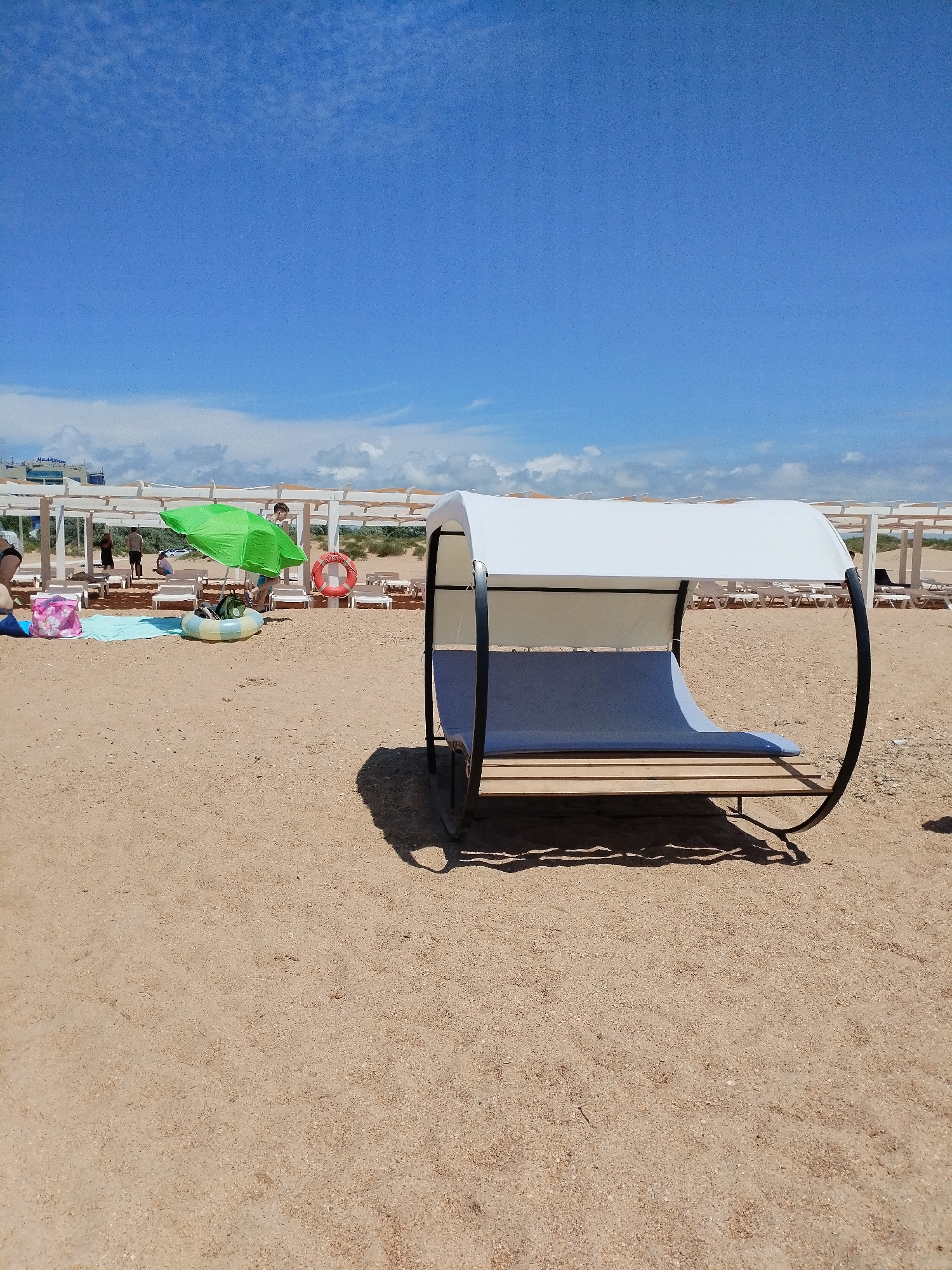 The image shows a sandy beach with several rows of white lounge chairs under a pergola structure. In the foreground, there is a modern white and blue shaded lounger with a curved metal frame. To the left, a green umbrella shades some inflatable toys, beach bags, and a life preserver ring. Further back, people are visible walking and sitting. In the distance, buildings and a cloudy blue sky are visible. The location is identified as Vinogradny, Russia.