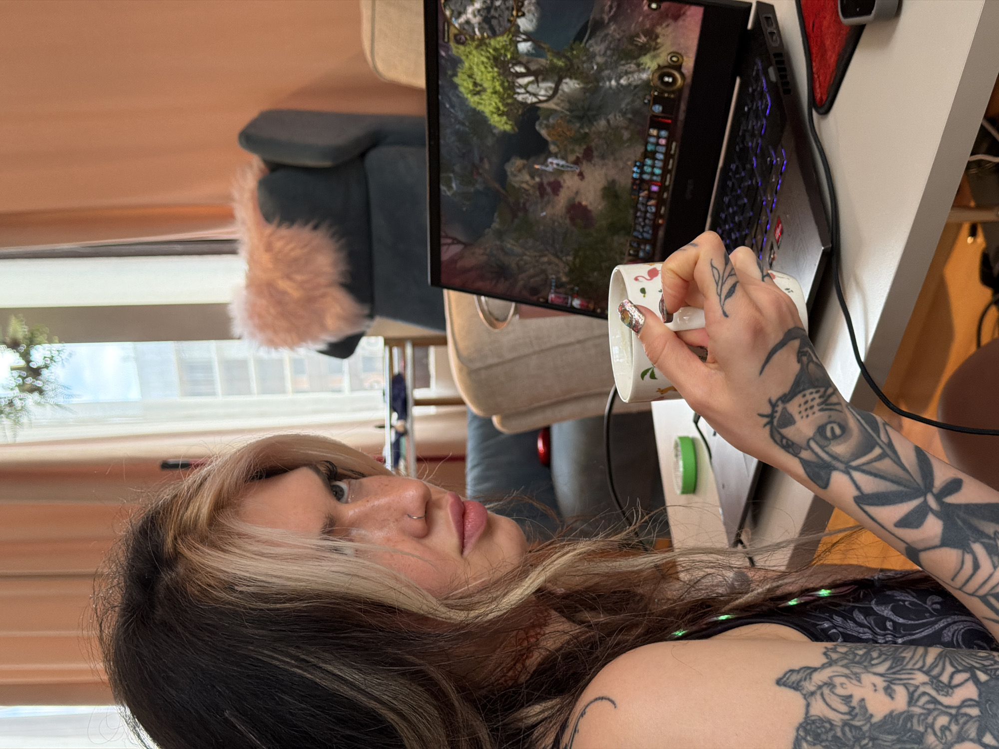 A person is holding a white mug with a flamingo design near a laptop. The screen shows a video game. The person has visible tattoos on their arm. The background shows a glimpse of a room with a window and furniture. The location is unspecified, but the prompt mentions Budva, Montenegro.