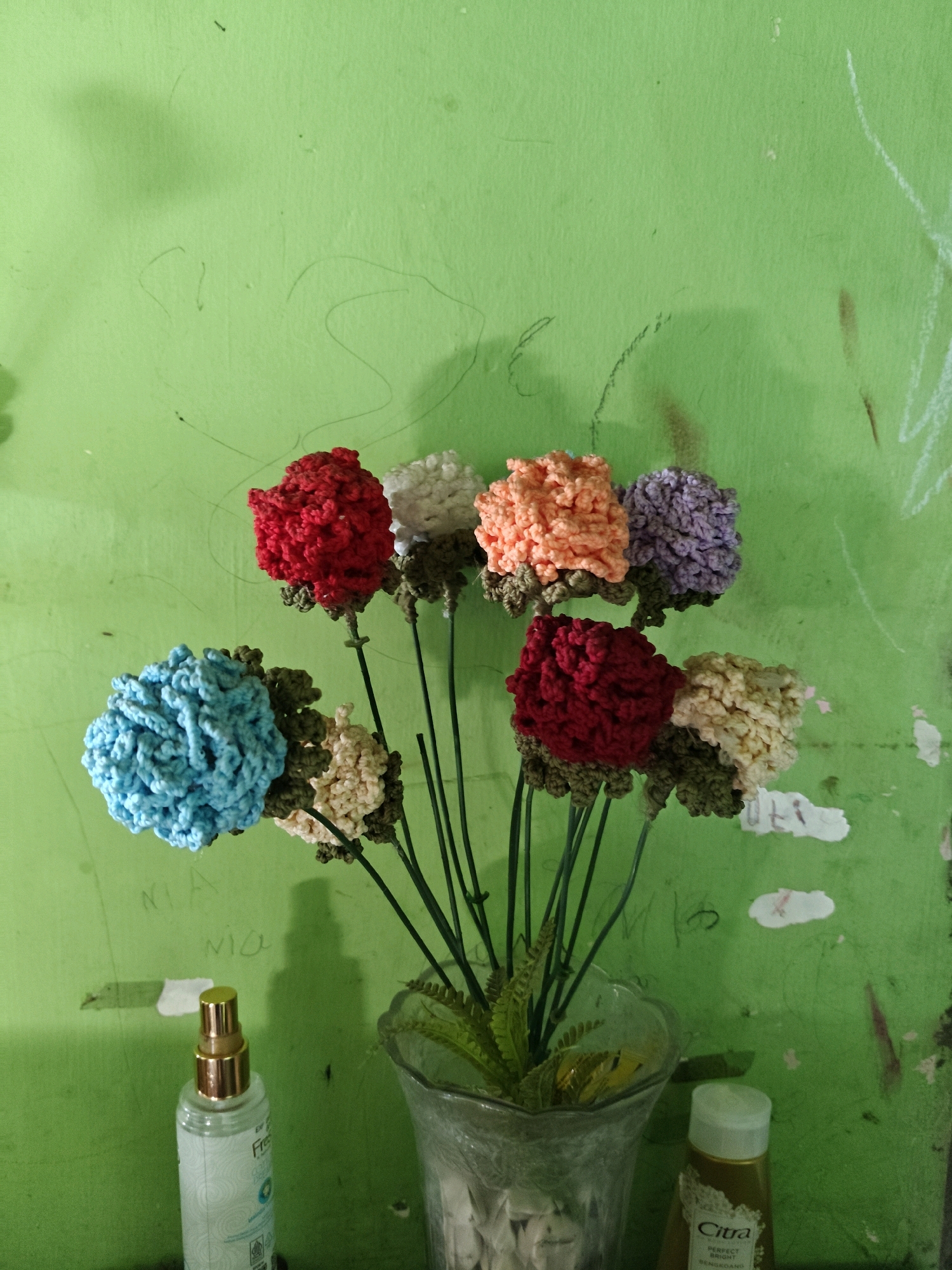 The image shows a bouquet of seven crocheted flowers in various colors, including red, white, orange, purple, light blue, and cream, arranged in a clear glass vase on a surface. The vase contains green artificial foliage. Behind the flowers, a green wall is visible with some scribbles and marks. In the foreground, to the left, is a clear spray bottle with a gold cap, and to the right, a small bottle with a white cap labeled "Citra." The overall scene appears to be indoors, likely in a residential setting in the city of Sumberwono, Indonesia, suggested by the presence of a product with the brand name "Citra," which is popular in Indonesia.