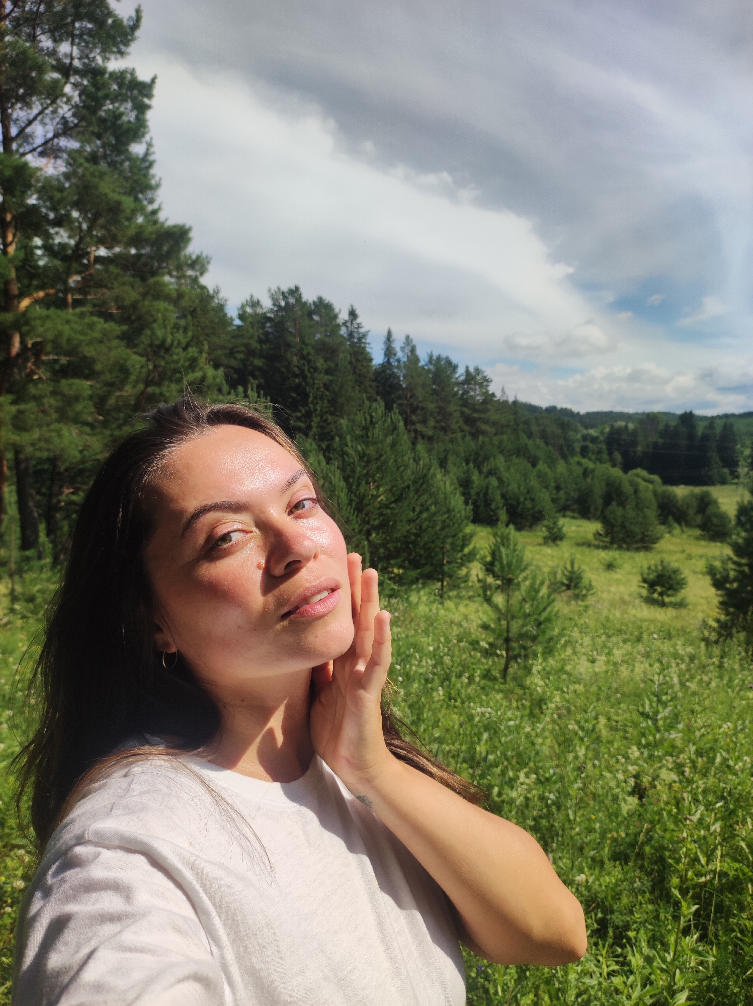 A close-up, waist-up selfie taken outdoors shows a young woman with dark hair, wearing a white t-shirt. She is touching her face with her right hand, her left arm is bent at the elbow and her hand is not visible. Her eyes are open and she is looking towards the camera. The background is a lush green landscape with rolling hills covered in trees and grass under a partly cloudy sky. The location is identified as Foky, Russia.