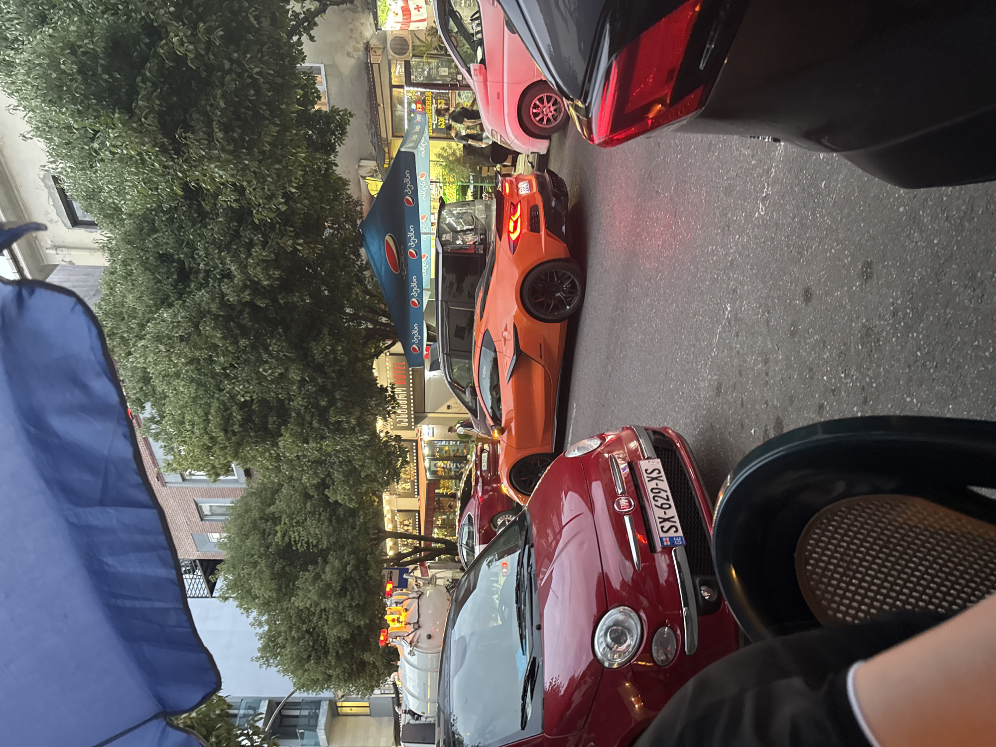 A street scene in Batumi, Georgia, shows several cars parked and in motion on a paved road. In the foreground, a red Fiat 500 is visible, with its license plate reading "SX-629-XS". To its right, an orange Chevrolet Corvette is parked, facing away from the camera. Behind the Corvette, a pink van is parked, and further back, a red sedan is also visible. Buildings line the street, with awnings and umbrellas suggesting commercial establishments. Trees with green foliage are interspersed among the buildings. The sky is partially obscured by a large blue umbrella in the upper left corner. The overall impression is that of a busy urban street on a daytime.