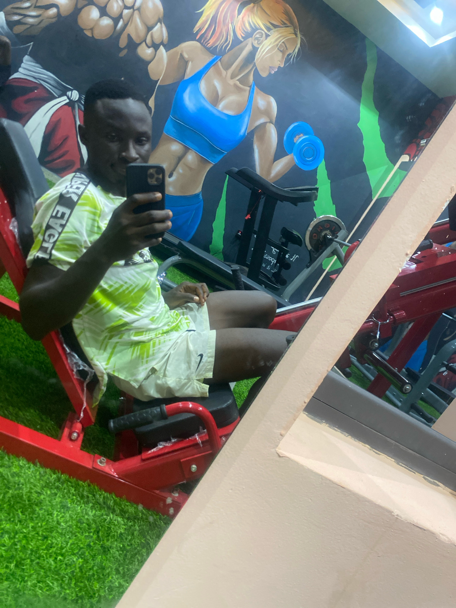 A young Black male is seated on a red exercise machine in a gym. He is holding a smartphone in his right hand, appearing to take a selfie. He is wearing a light green and white patterned t-shirt with black and white text on the sleeves, and light-colored shorts. The gym features a mural on the back wall depicting muscular figures and a woman lifting weights. Artificial grass covers the floor around the exercise equipment. The gym appears to be located in Barden Yana, Nigeria.