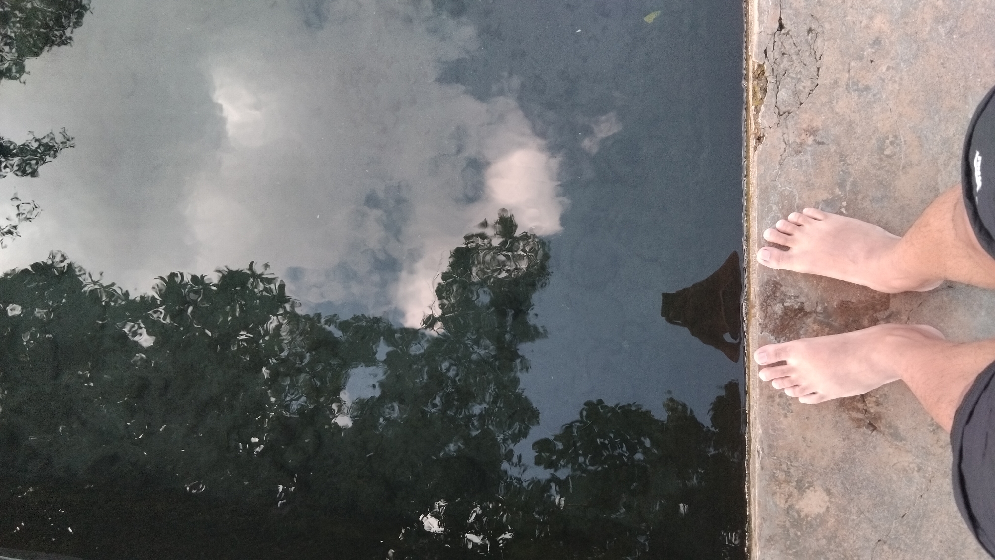 A first-person perspective shot shows a person's bare feet standing on a concrete ledge next to a body of dark water. The water reflects trees and clouds. The concrete ledge is weathered and cracked. The scene appears to be outdoors, possibly in a park or recreational area. The location is identified as Ngaglik, Indonesia.
