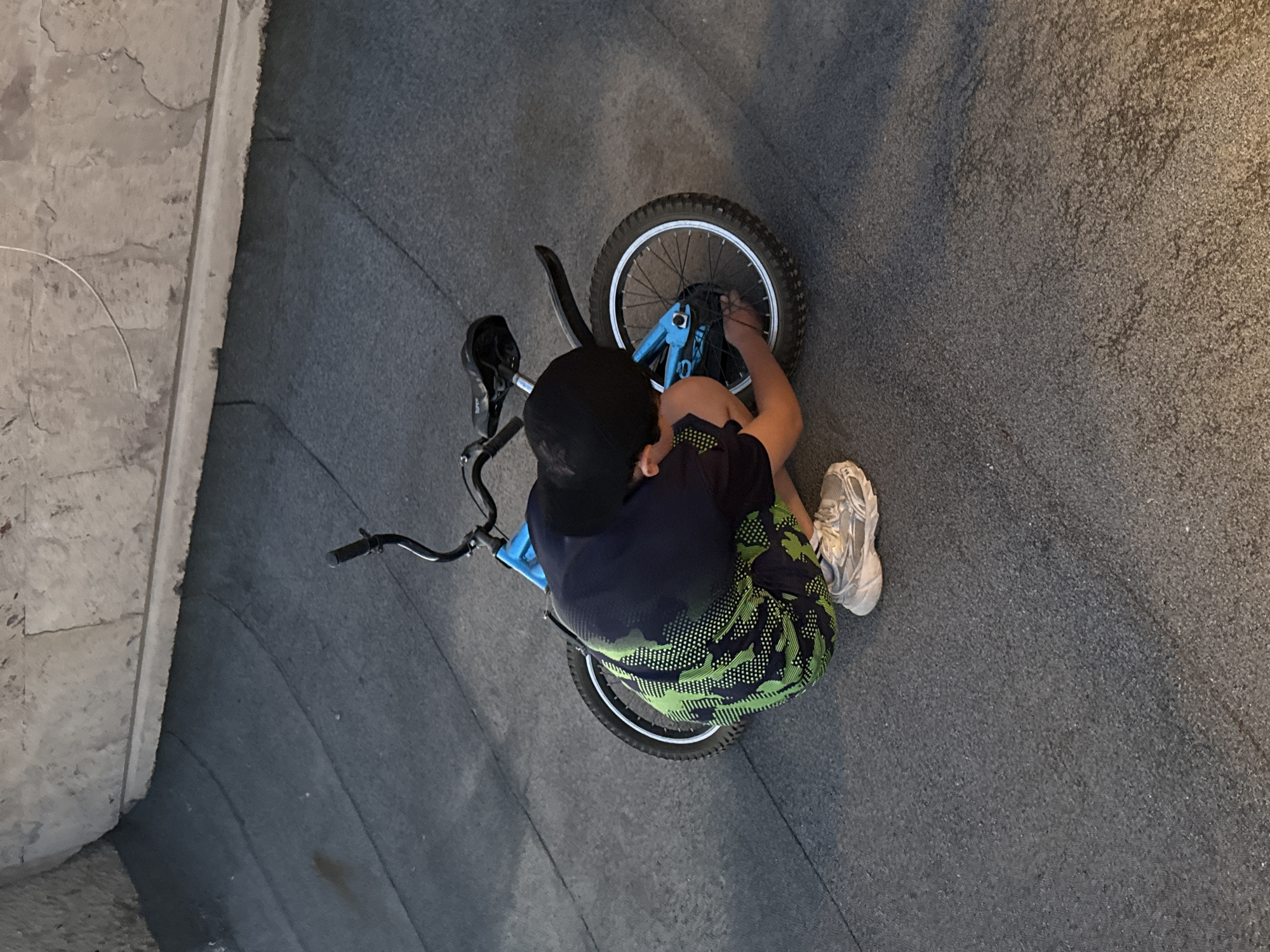 In Yerevan, Armenia, an overhead shot captures a sole individual, presumably male, crouching on a dark, granular paved surface. The person wears a black baseball cap, a dark t-shirt, and green and black patterned shorts, complemented by light grey athletic shoes. A blue child's bicycle lies horizontally on its left side directly in front of the individual, who is actively engaged with its rear wheel, with hands positioned near the hub and spokes. The background features a textured light grey concrete or stone wall with a distinct horizontal ledge running across its upper section.
