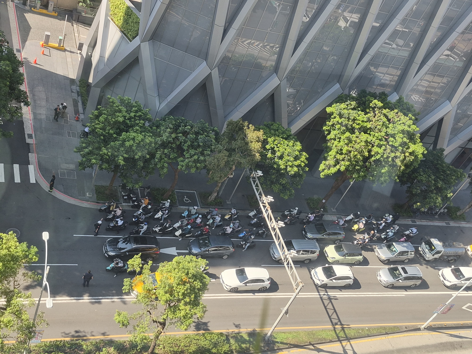 This aerial, wide-angle, eye-level shot depicts a street scene in Taipei, Taiwan, during daylight. The image is dominated by a modern, angular building with a glass facade and metallic structural elements on the right side, reflecting the sky and surrounding environment. A mature, leafy tree stands prominently in the foreground on the left, with more trees and greenery lining the street.
The street itself is a multi-lane road with visible lane markings. A significant number of parked and moving motorcycles, scooters, and a few cars are present. On the left side of the street, several people are visible, including individuals standing on the sidewalk and near parked vehicles. One person in dark clothing is walking on the sidewalk. A black SUV is parked in a lane of traffic. Further down the road, a yellow taxi is partially visible.
On the right side of the street, a white car is moving, and a silver SUV is parked. Several other cars and motorcycles are scattered throughout the scene, with some individuals appearing to be riding or standing near them. A traffic light pole with multiple security cameras is positioned in the middle of the road. The scene suggests a typical urban environment with moderate traffic and pedestrian activity.