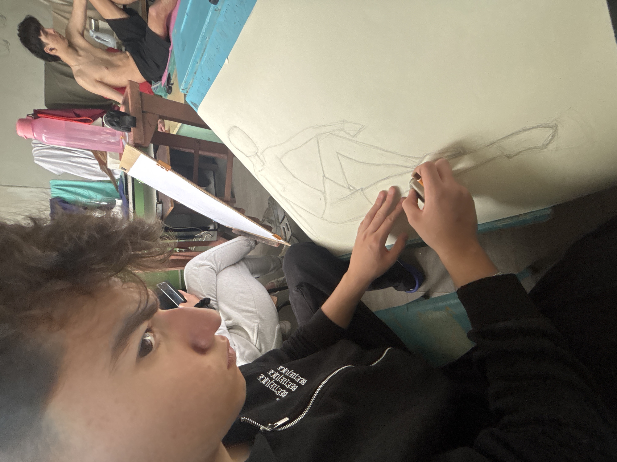 The image depicts an indoor art session, likely in an art studio or informal setting in Troyan, Bulgaria. In the foreground, partially visible, an individual with brown, curly hair is focused on drawing. This artist is wearing a black zippered jacket with the word "BEETLEJUICE" repeated multiple times in white capital letters. Their hands are shown actively working on a large, light-colored drawing board, using an eraser or similar tool to refine a pencil sketch of a reclining human figure.
In the background, another individual is posing as the model for the drawing. This person is shirtless, wearing black shorts, and is lying down on a surface, appearing relaxed. Various personal items are visible around the model, including a pink water bottle, folded fabrics, and wooden furniture components. A white drawing board or sheet of paper is also visible on an easel or stand positioned between the artist and the model. The scene is well-lit, suggesting either artificial lighting or bright, diffused natural light from an unseen window. The overall ambiance is one of focused artistic creation.