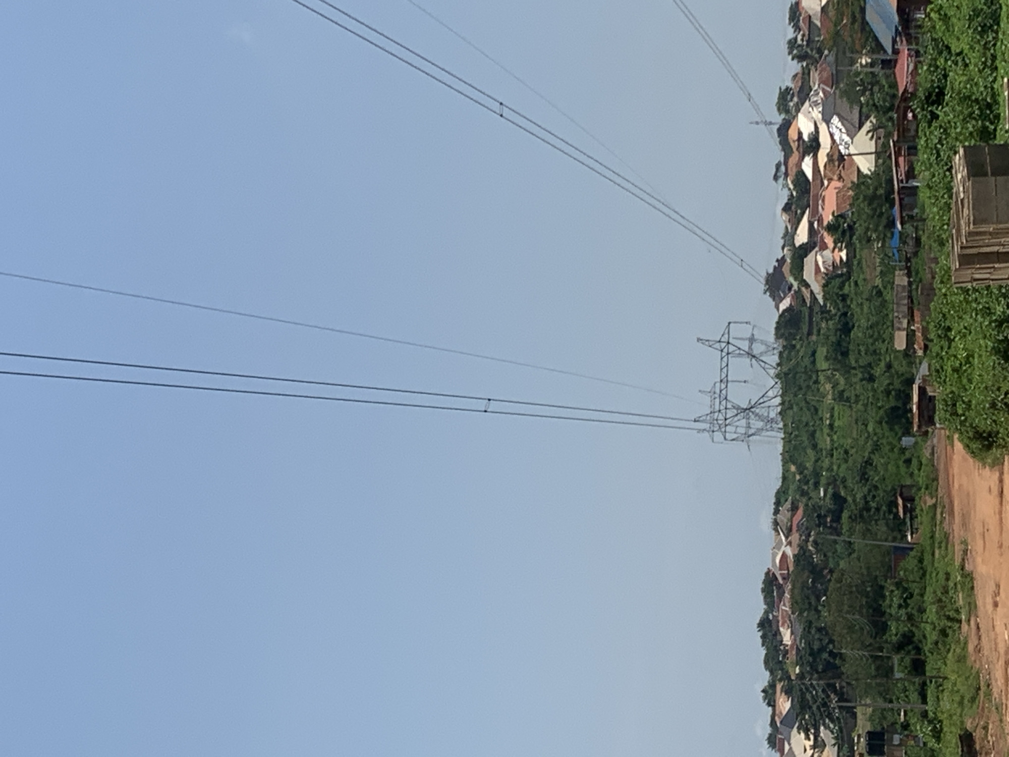A vertical, eye-level, full shot captures a rural landscape under a clear sky. The scene is dominated by tall electricity pylons and their overhead cables, which stretch across the frame. Below these pylons, a densely populated area of houses with varied roof colors and styles is nestled amongst lush green trees and vegetation. A dirt road runs through the foreground, bordered by more greenery. The image appears to be taken in a suburban or semi-rural part of the city of Unknown, Unknown.