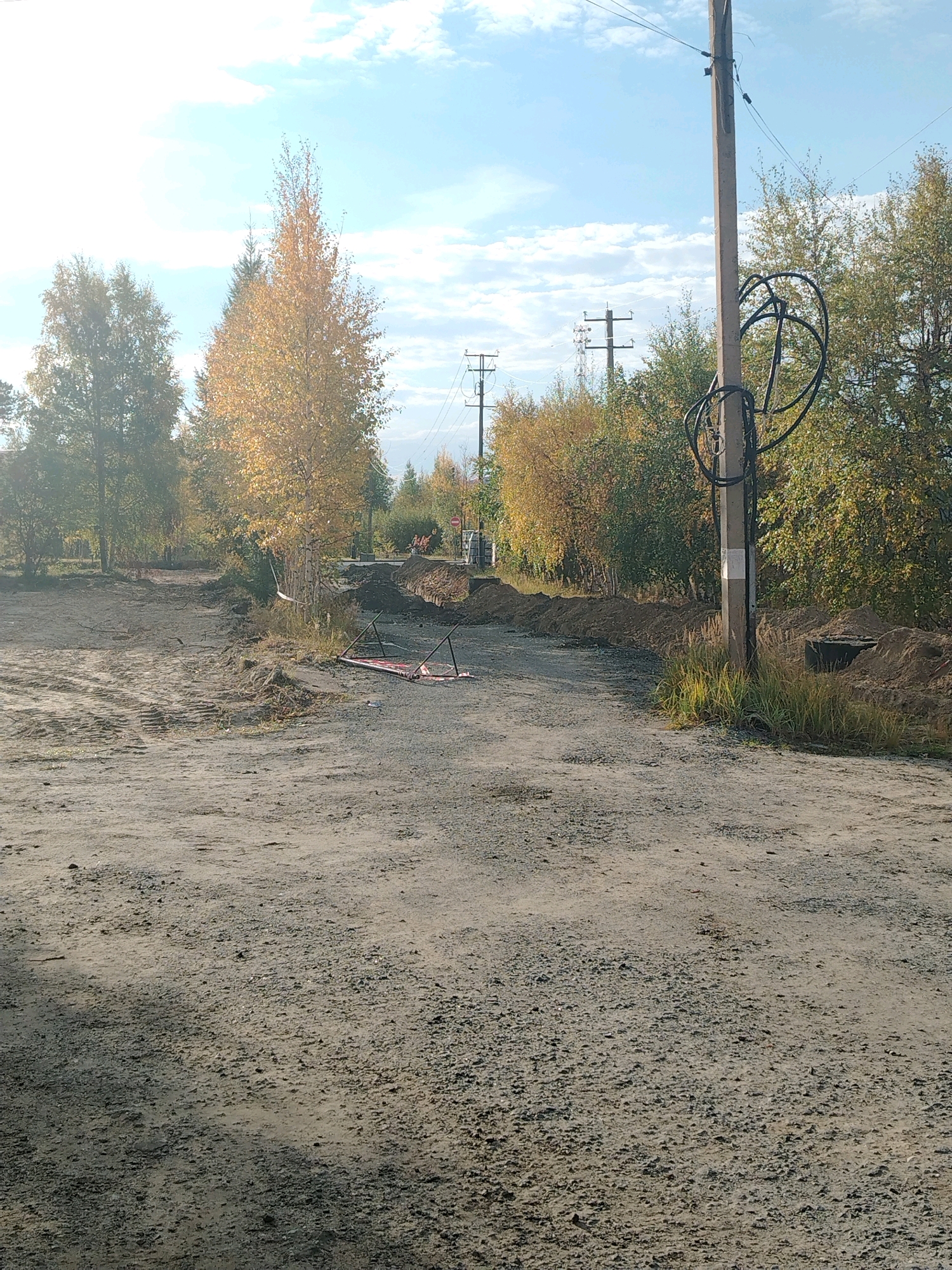 This is a forensic, matter-of-fact description of an outdoor scene in Muravlenko, Russia. The image depicts a dirt road with piles of excavated earth along its sides, suggesting ongoing construction or utility work. Several trees with autumn foliage line the background, some with golden leaves, indicating the season. A concrete utility pole stands prominently on the right side of the road, with coiled black cables wrapped around its base and electrical wires extending upwards. In the middle ground, a red and metal frame object lies on the ground. In the distance, more utility poles and trees are visible under a bright sky with scattered clouds. The ground is covered in loose gravel and dirt. There are no people or animals visible in the image.