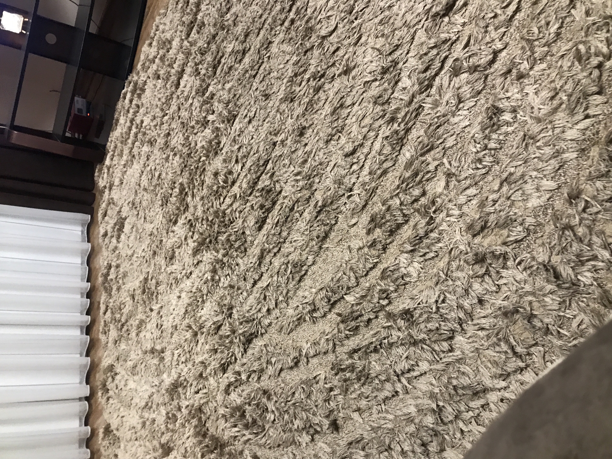 The image depicts a close-up view of a textured, beige or light gray shag rug. The rug's fibers are densely packed and appear to be somewhat worn or flattened in certain areas. To the left of the rug is a portion of white, horizontal blinds. In the upper left corner, a dark-colored, partially visible shelving unit or cabinet is present. The scene does not provide information about the location in the city of Yerevan, Armenia.