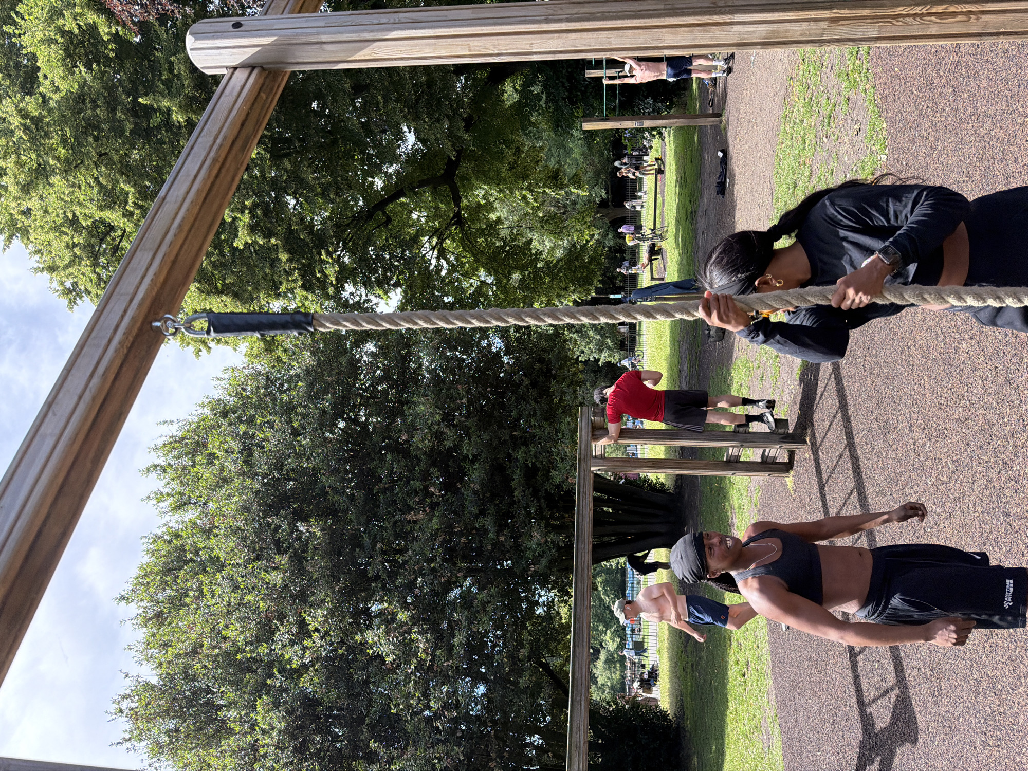 In a park in London, United Kingdom, on a sunny day, two women are about to climb a rope hanging from an outdoor fitness structure. The woman in the foreground on the right is wearing a black long-sleeved top and black pants and is holding onto the rope. The woman on the left is wearing a black sports bra and black shorts and has a bandana on her head. Several other people are in the background engaged in various activities like exercising and sitting on benches. The fitness equipment is made of wood and metal, and the ground is covered in wood chips. Lush green trees surround the area, indicating a park setting.