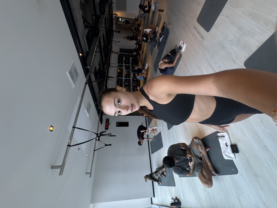 This image depicts an indoor fitness studio located in Bangkok, Thailand, identified by the digital clock displaying "Bangkok" on the wall. In the foreground, a young adult female with dark hair tied back, wearing a black sports bra and black athletic shorts, captures a selfie, looking directly at the camera with a neutral expression.
The background and mid-ground reveal approximately 15-20 individuals participating in a class, likely yoga or stretching, spread across a light wooden floor on individual grey yoga mats. Participants exhibit various postures: some are seated upright, some are performing forward folds or other stretches, and others are observing their mobile phones. Notable individuals include a male in the mid-left foreground seated cross-legged on a mat, examining his phone, with visible tattoos on his legs. Another person on the far left wears a green cap and camouflage trousers, lying on a mat.
The environment features white walls, a large window on the left side revealing an external building, and a ceiling with exposed dark metal framework, recessed lighting, and square air conditioning vents. Metal bars with black hanging straps are suspended from the ceiling. A digital clock on the back wall displays "10:58:11" with the city "Bangkok" printed beneath it. Shelving units in the far-right background hold numerous black spherical objects, possibly medicine balls or weights. Personal items such as water bottles, small white towels, and mobile phones are visible near several mats.