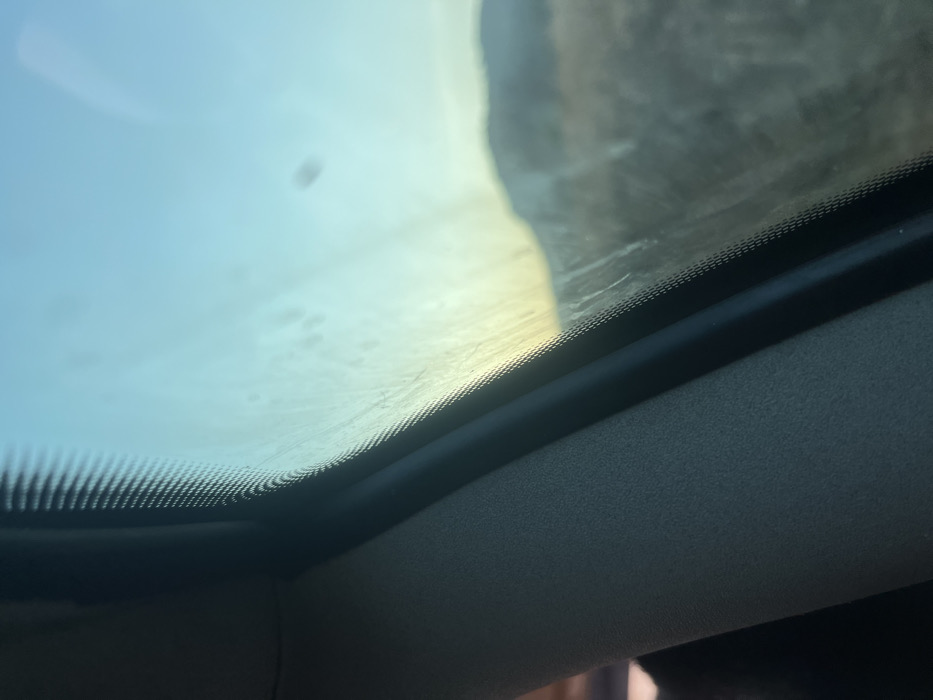 Looking upwards, the image captures the interior of a vehicle. A clear, seemingly clean sunroof is visible, with a blurry outside scene partially visible. The interior trim of the car is visible, consisting of a dark rim that goes around the sun roof and a lighter-colored upholstered roof.