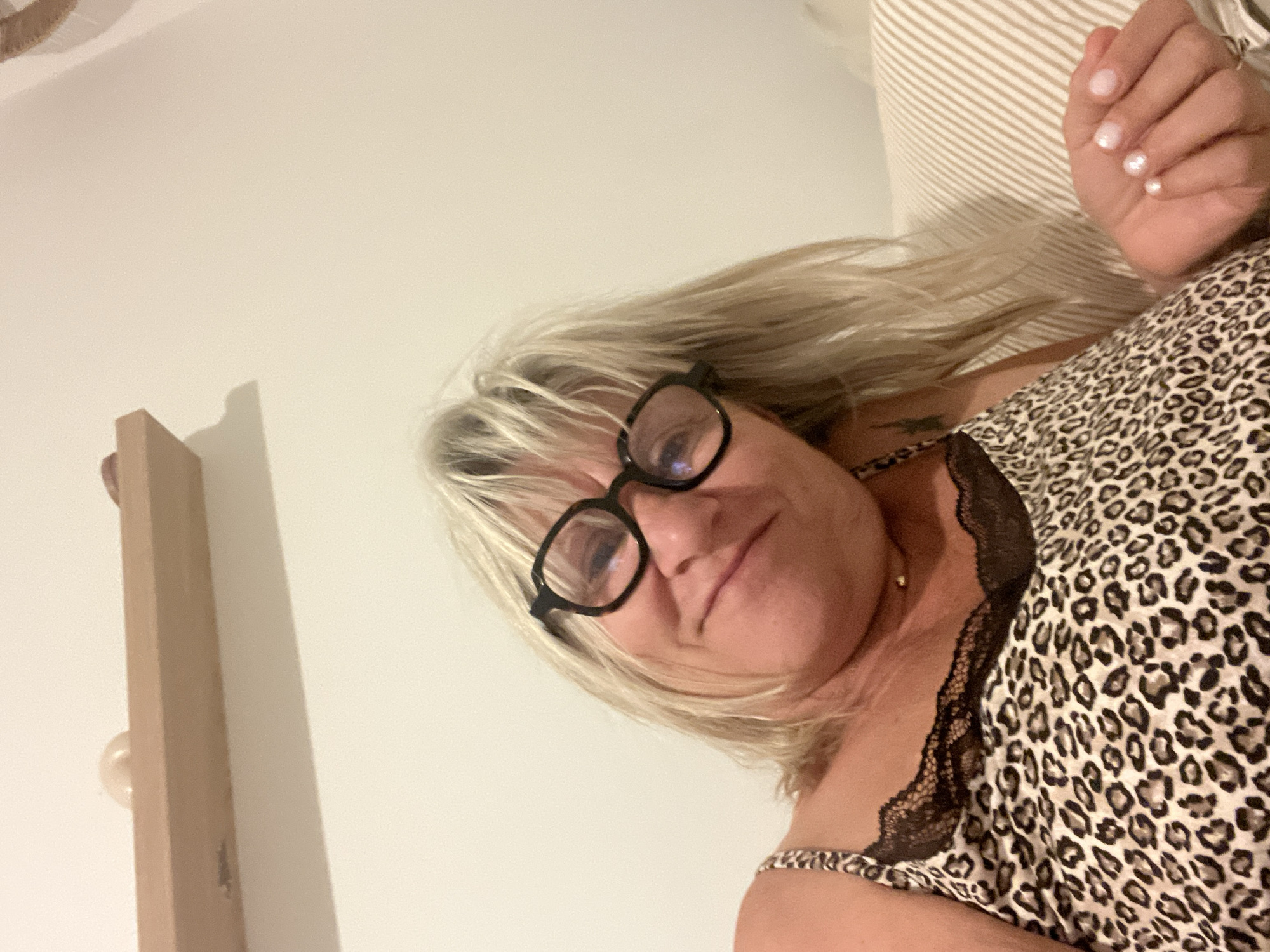 A blonde woman wearing glasses and a leopard-print camisole is lying on a striped sofa. She is holding up her left hand, showing off her fingernails, which are painted a light pink and appear to have iridescent accents. The background is a plain, off-white wall, with a wooden shelf mounted horizontally above the woman's head. There are no discernible elements in the image that would indicate a location in Cannes, France.