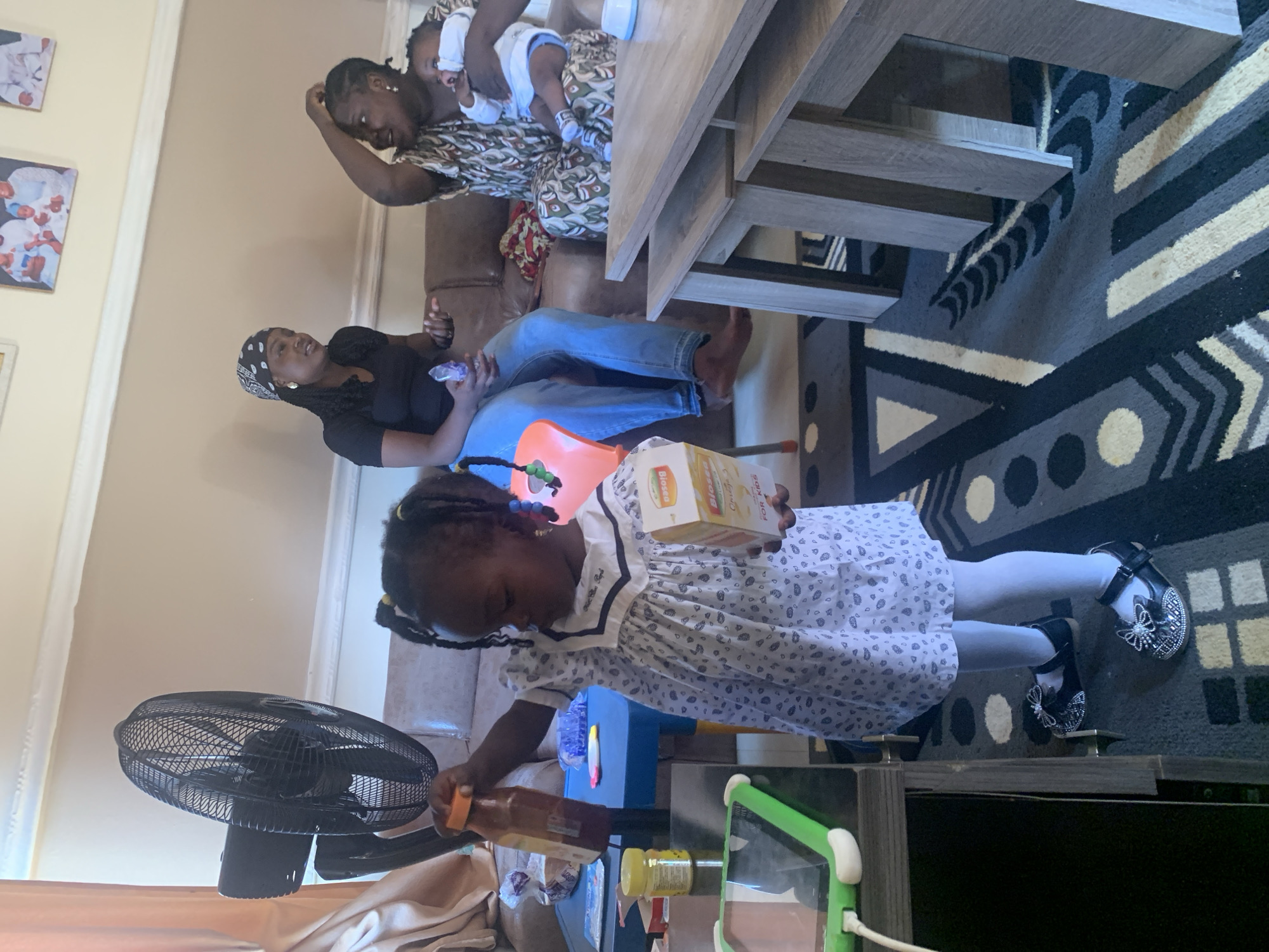 In Gura Top, Nigeria, a young girl wearing a patterned dress and white tights stands in a living room, holding a bottle and a box. To her left, a black floor fan is visible. Behind her, two women are seated on a brown couch. One woman, wearing a black t-shirt and a patterned headscarf, gestures with her hands. The other woman, in a patterned dress, holds a baby. A wooden coffee table with nested tables is positioned in the foreground. The room is decorated with framed pictures on the wall and a patterned rug on the floor.