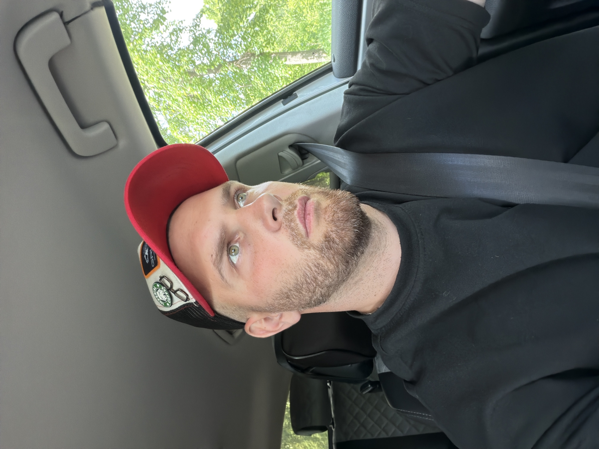 A man is depicted from a low-angle perspective, seated inside a car on a bright, sunny day in Chaikovsky, Russia. The image is oriented sideways, but if rotated correctly, the man is looking upwards and slightly to his right, with a neutral and observant expression. He has green eyes, a light brown beard, and fair skin. He is wearing a black long-sleeved hoodie or sweatshirt. On his head, he wears a red baseball cap with a black mesh back. Visible patches on the cap include one with white and green elements that reads "IMPERIAL COFFEE" and another partially visible patch starting with "GR". A small metallic pin or emblem is attached to the "IMPERIAL COFFEE" patch. He is wearing a black seatbelt across his chest. The car's interior features a grey headliner and grab handle, and a quilted pattern is visible on the seat or headrest. Through the car window, vibrant green foliage from trees can be seen, indicating an outdoor setting during daylight.