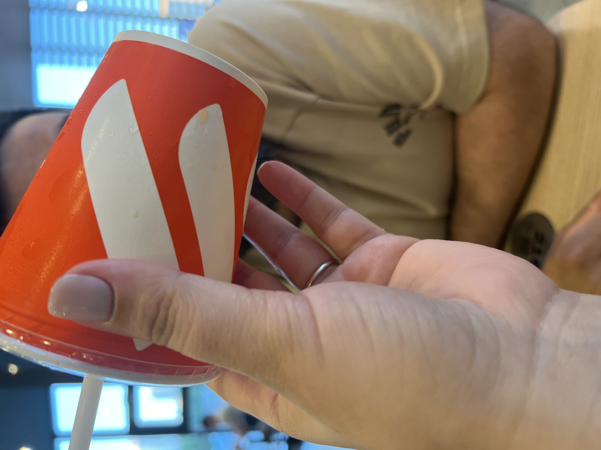 A close-up shot shows a hand holding an orange and white paper cup with a straw. The cup is tilted, and condensation is visible on its surface. The hand has a light-colored nail polish and is wearing a silver ring on the ring finger. In the background, a person in a light-colored t-shirt is seated, with their arms resting on a table. The environment appears to be indoors, possibly a cafe or restaurant, with blurred windows and interior elements suggesting a public space. The image does not contain enough visual information to determine its specific location in Yekaterinburg, Russia, or any specific actions or interactions beyond the act of holding the cup.