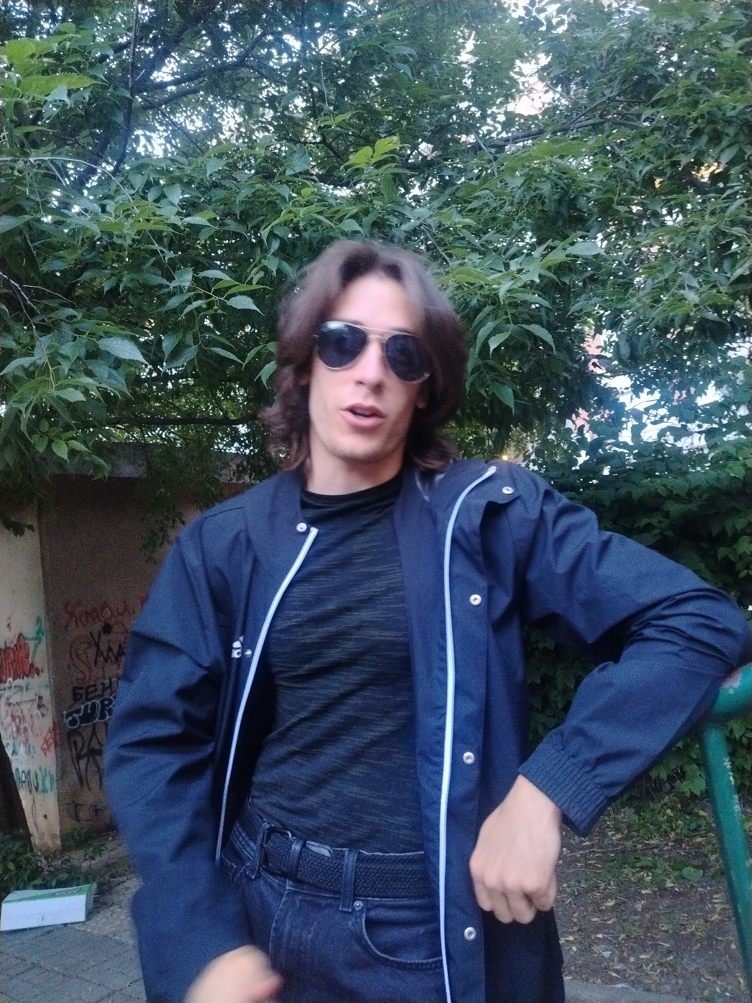 A man stands outdoors, posing in front of a lush green tree. He wears sunglasses, a black t-shirt, a dark blue jacket with white piping, and dark jeans. He is leaning his arm on a green rail. Behind him and to his left is a wall with graffiti. The location is likely in an urban setting in Burgas, Bulgaria.