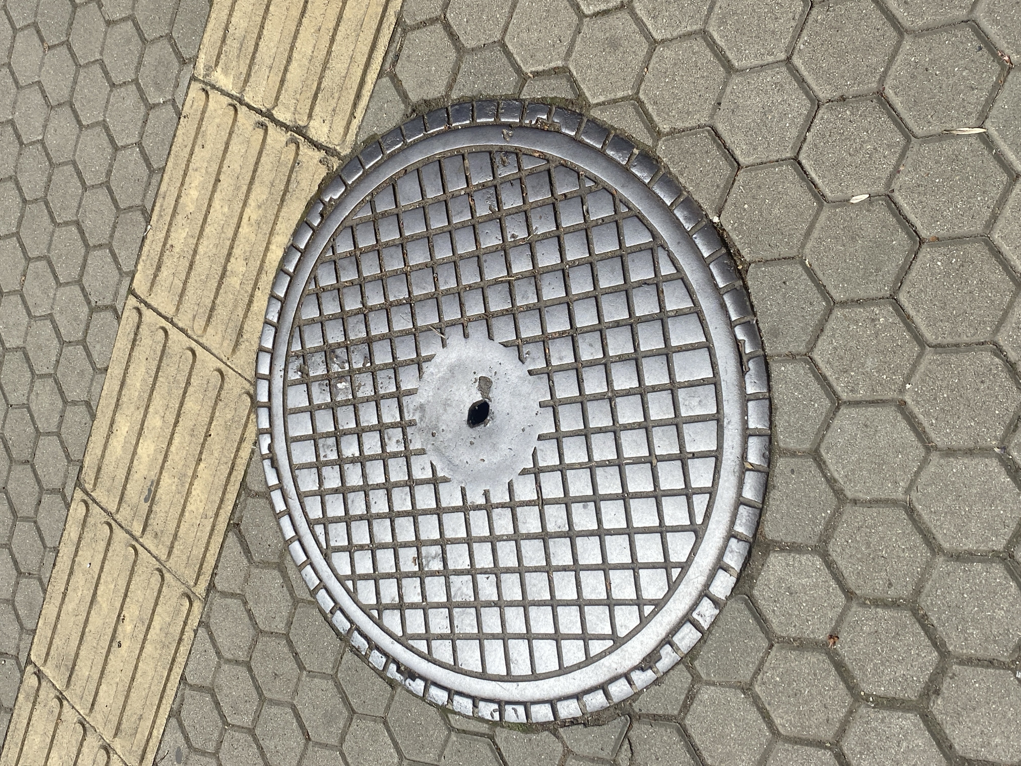 The image shows a circular metal manhole cover with a grid pattern, set in a pavement composed of hexagonal concrete pavers. A textured yellow strip, likely tactile paving for the visually impaired, runs along the left edge of the frame. The manhole cover is slightly off-center, positioned towards the middle of the image. There's a discolored, splattered area around the center of the cover. The pavers are light gray and show signs of wear and minor debris. The location is potentially within the city of Sofia, Bulgaria based on the user's prompt. There is no evidence of any people in this image.