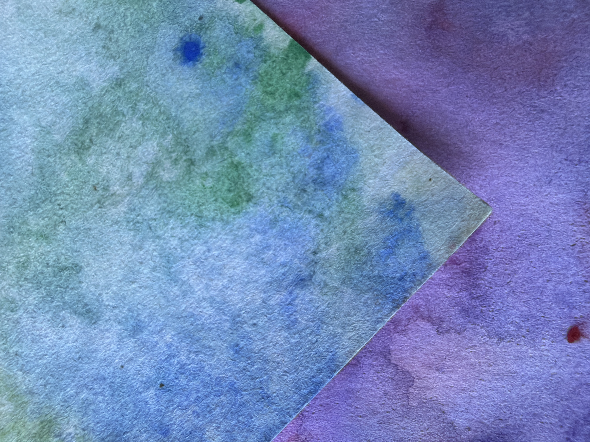 The image displays a close-up, abstract view of two pieces of textured paper with watercolor paint. One piece, in the foreground, is diagonally oriented and shows washes of blue and green. The other piece, underneath and to the left, is predominantly purple with some pinkish hues. There is a small, intense blue blotch on the upper right portion of the blue and green paper, and a dark speck near the center. The texture of the paper is rough, with visible fibers. There are no people, recognizable landmarks, or context suggesting a specific location within Ufa, Russia.
