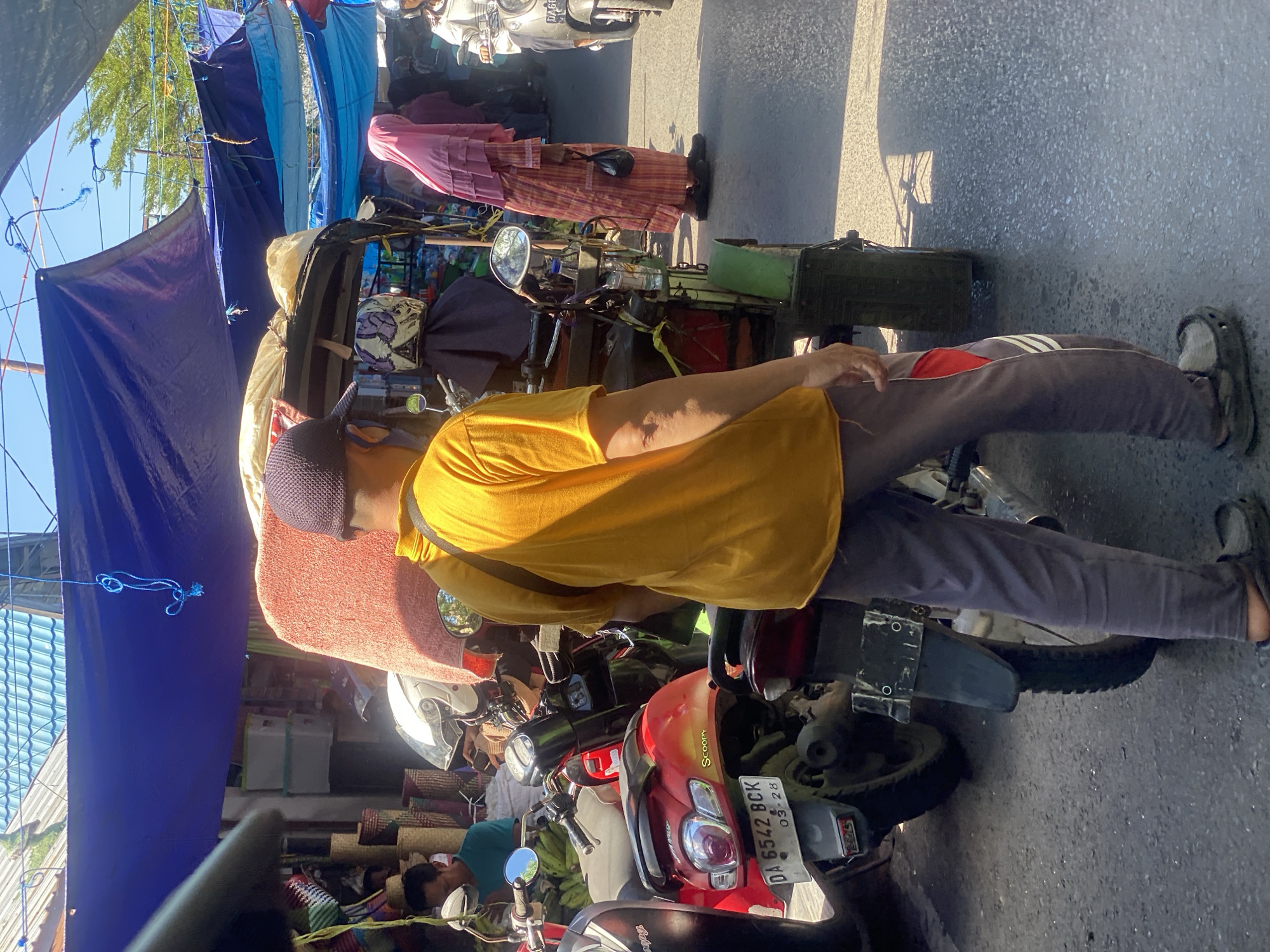 The image captures a street scene in Desa Tunggul Irang, Indonesia, under a clear sky. The foreground is dominated by a man wearing a yellow t-shirt, dark pants, and sandals, standing with his back to the camera, facing a three-wheeled vehicle that appears to be a modified motorcycle with a passenger enclosure covered by a pink blanket. To the left of the man, a red scooter is parked, displaying the license plate "DA 6542 BCK 03-28".
Further into the scene, a woman in a pink headscarf and patterned dress is visible, carrying a black plastic bag. Other motorcycles are parked or in motion, and various market stalls with blue awnings are set up along the street. Baskets and other goods are displayed in the background. The ground is paved asphalt. The overall impression is of a busy marketplace or thoroughfare.