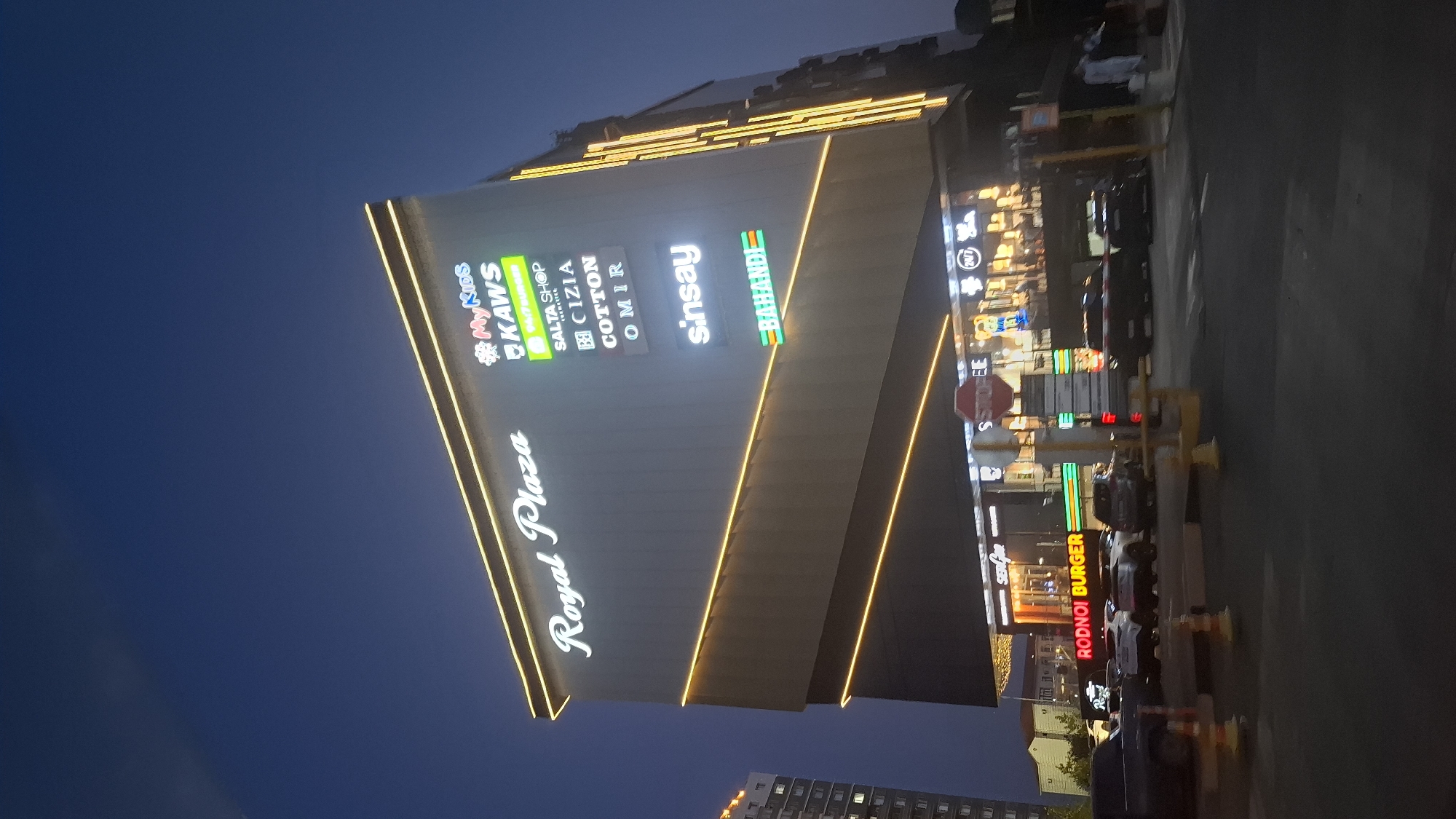 The image shows the exterior of the Royal Plaza shopping center at dusk in Shymkent, Kazakhstan. The building is modern with prominent signage. "Royal Plaza" is displayed in large white script on the upper facade. Below this, a vertical array of smaller illuminated signs advertises various brands including "MyKids," "KAWS," "SALTA SHOP," "CIZIA," "COTTON," "OMIR," "sinsay," and "BAHANDI." The building's exterior is illuminated by warm yellow accent lighting that follows its geometric lines.
At the ground level, illuminated signs for "RODNOI BURGER" are visible, along with other storefronts that are less clear due to lighting. A red stop sign is mounted on a pole in front of the building. Several cars are parked or driving in the foreground, and some traffic barriers are present. People are visible in the background on the right side of the image, walking near the entrance of the shopping center. The sky is a deep blue, indicating twilight. A tall apartment building is visible to the far left of the frame. The scene suggests a busy commercial area in an urban setting.