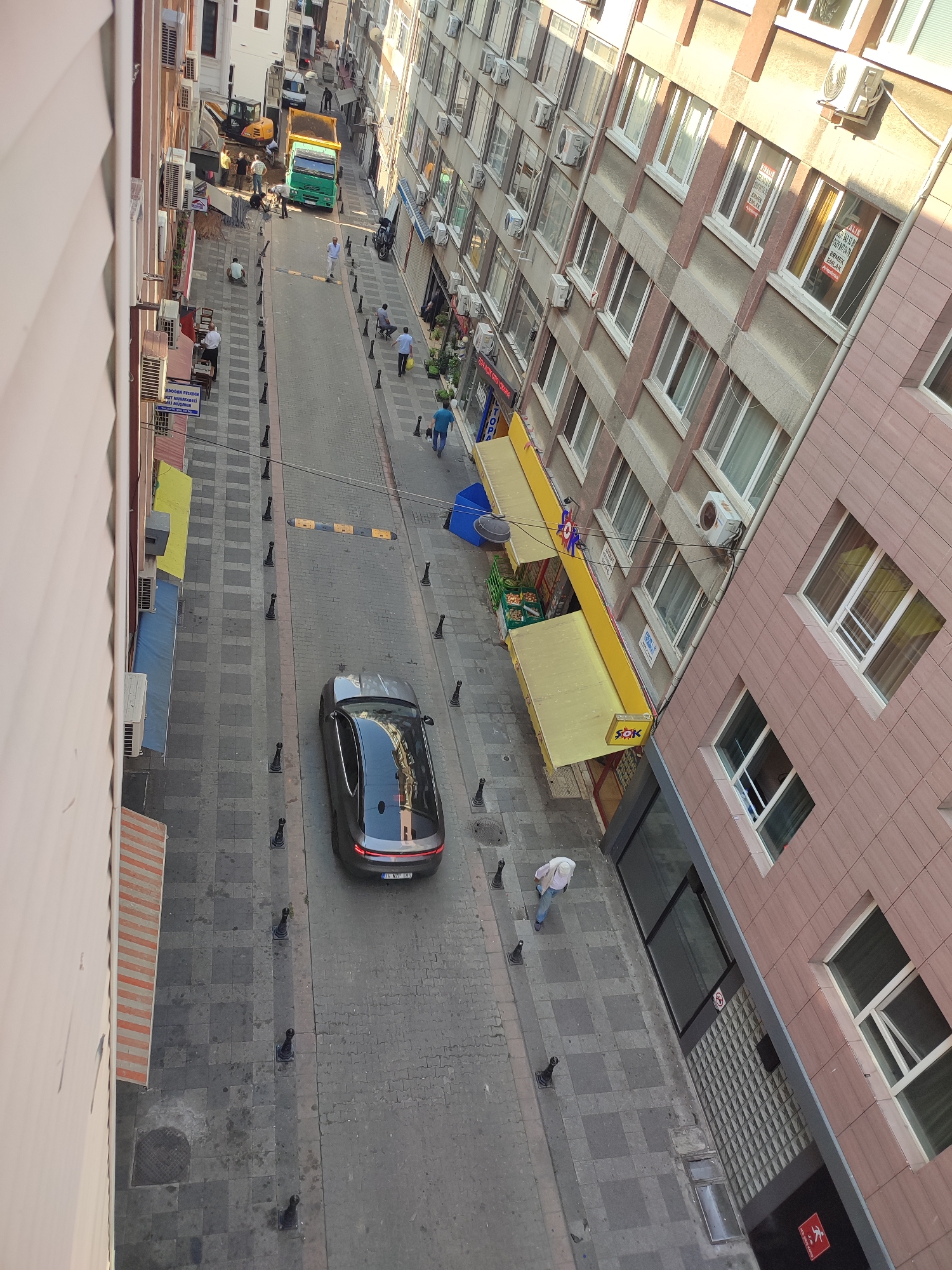 An aerial shot depicts a narrow city street in Kadıköy, Turkey, lined with multi-story buildings. Construction is underway in the upper left of the image, with an excavator and a dump truck present. Several construction workers are visible near the machinery.
Further down the street, a black sedan is parked in the lane. People are walking on the sidewalks, some near shops with awnings. The buildings have numerous windows, and air conditioning units are mounted on their facades. Streetlights and bollards line the sidewalks. The ground is paved with a mix of brick and stone. The overall scene suggests a busy urban environment with ongoing construction and daily life.