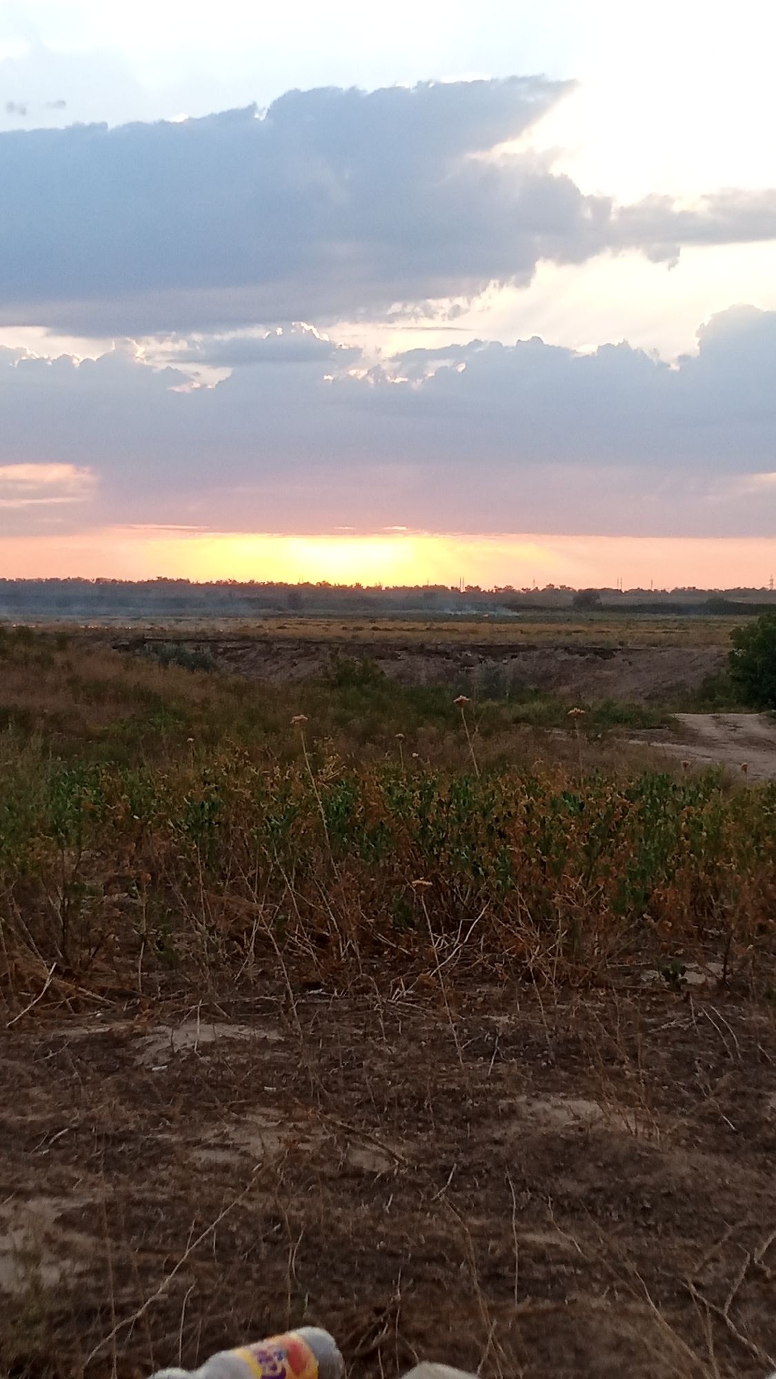 The image shows a wide-angle view of a rural landscape at sunset, likely in Novyy Put, Kazakhstan. The sky is filled with dramatic clouds, illuminated by the setting sun which casts a warm glow across the horizon. Rays of light pierce through the clouds, creating a celestial spectacle. Below the sky, a vast expanse of dry, grassy terrain stretches out, with some sparse vegetation in the foreground. In the distance, a tree line marks the edge of the landscape, and faint silhouettes of structures suggest the presence of a settlement. The ground is covered in dry earth and withered plants, indicating an arid environment. The overall impression is one of natural beauty and tranquility, with a hint of the vastness of the steppes.