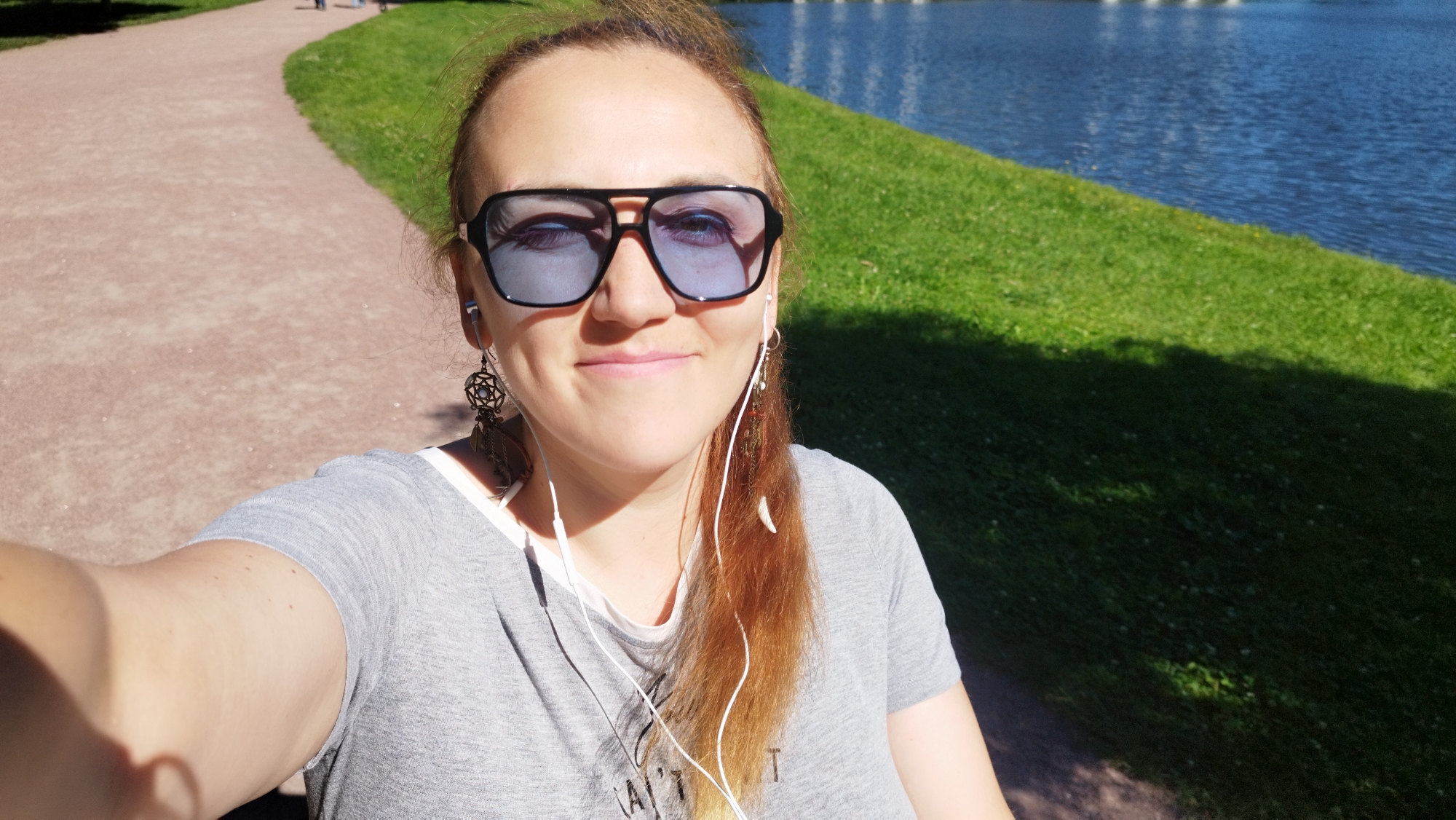 A woman wearing sunglasses and headphones takes a selfie in a park. She is wearing a grey t-shirt and has red hair. In the background, there is a path, a body of water, and green grass. The image appears to be taken in the Catherine Park in Pushkin, a suburb of Saint Petersburg, Russia, due to the architectural style of the path and the presence of a pond, characteristic of parks in the area. The time of day appears to be daytime, with bright sunlight casting shadows.