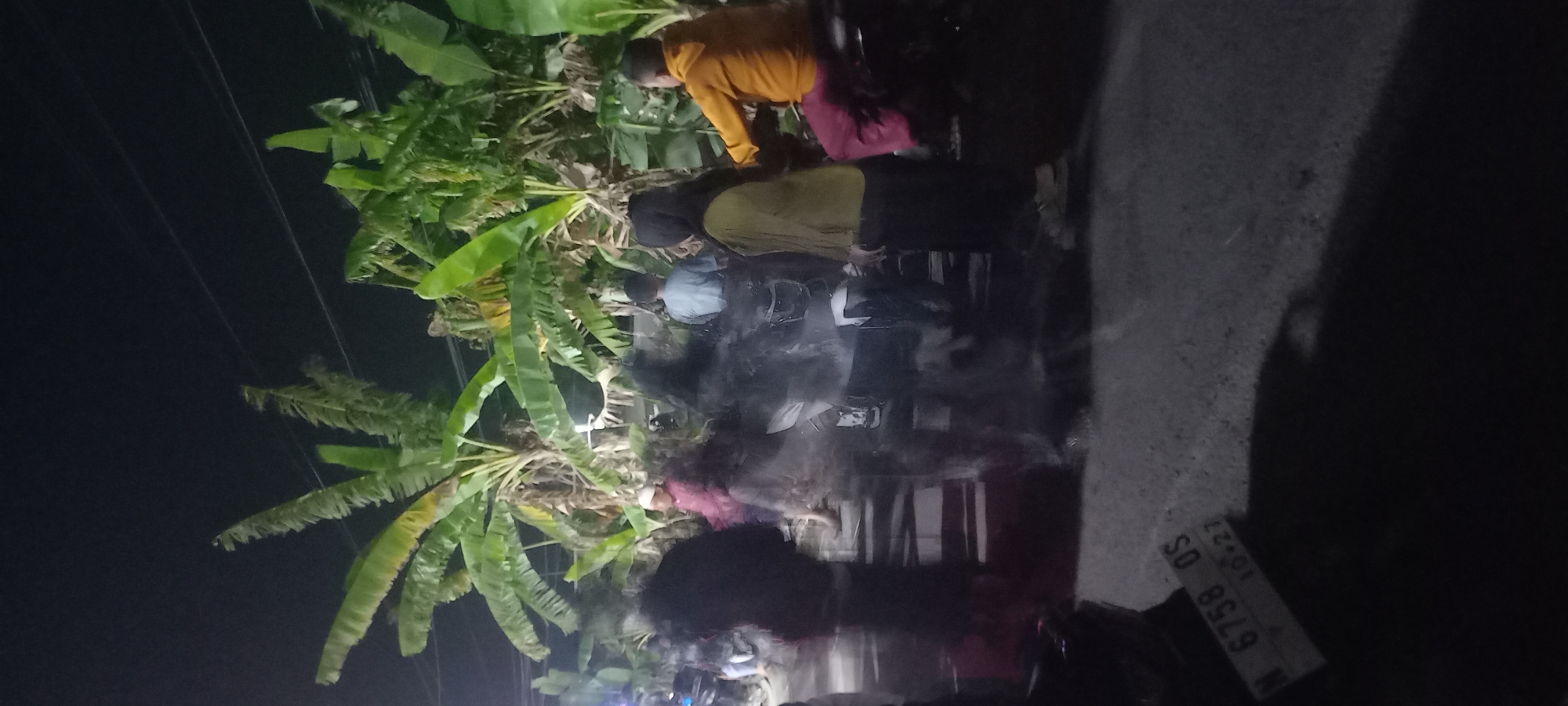The image, taken at night in Tongaswetan, Indonesia, depicts a street scene with several individuals and motorcycles. The foreground shows a license plate reading "N 6758 OS 10.27". In the middle ground, motion blur obscures the details of several people, some of whom appear to be near motorcycles. Further back, banana trees line the area, and additional figures are visible, including one person in a bright orange jacket and another in a yellow top. The overall impression is of a gathering or activity occurring on a street at night.
