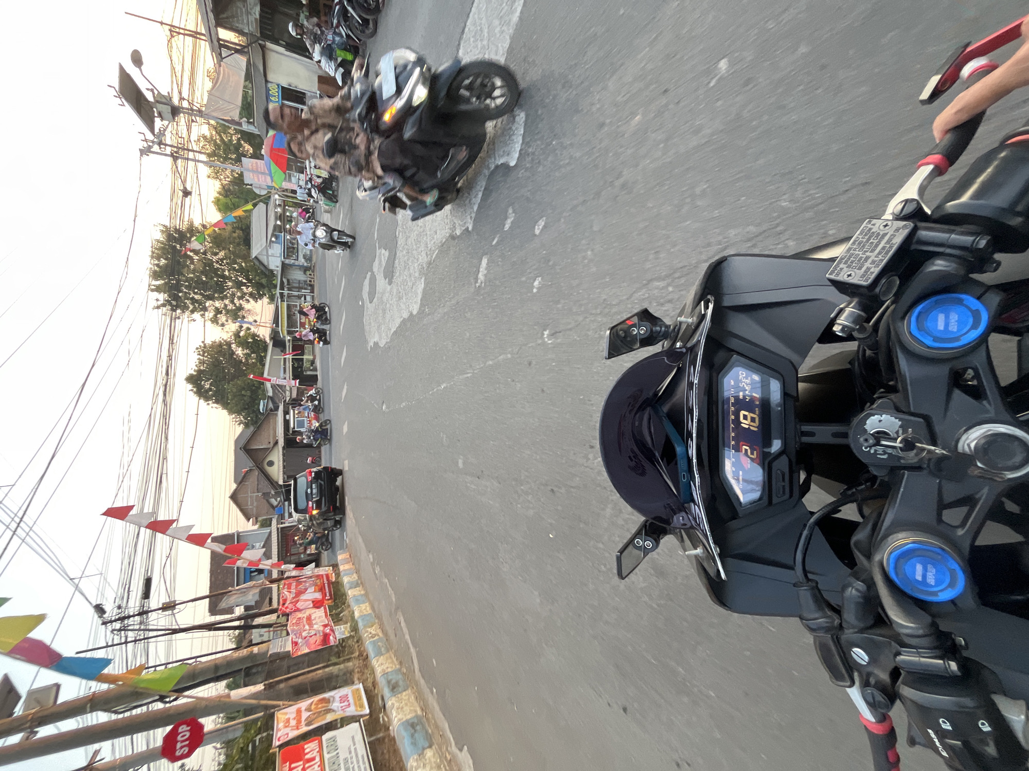 The image depicts a street scene in Pelang, Indonesia, taken from the perspective of someone riding a motorcycle. The road is paved and appears to be a two-lane street with moderate traffic. Several motorcycles and at least one car are visible on the road. The motorcycle in the foreground has a digital display showing "2 18" and other indicators. Red and white Indonesian flags are hung along the roadside, suggesting a festive or commemorative occasion. Various advertisements and signs are also present on poles and buildings lining the street. The architecture suggests a mix of residential and commercial buildings. The sky is clear with the sun appearing to be setting or rising, casting a warm light. The overall environment is that of a typical street in a populated area of Indonesia.