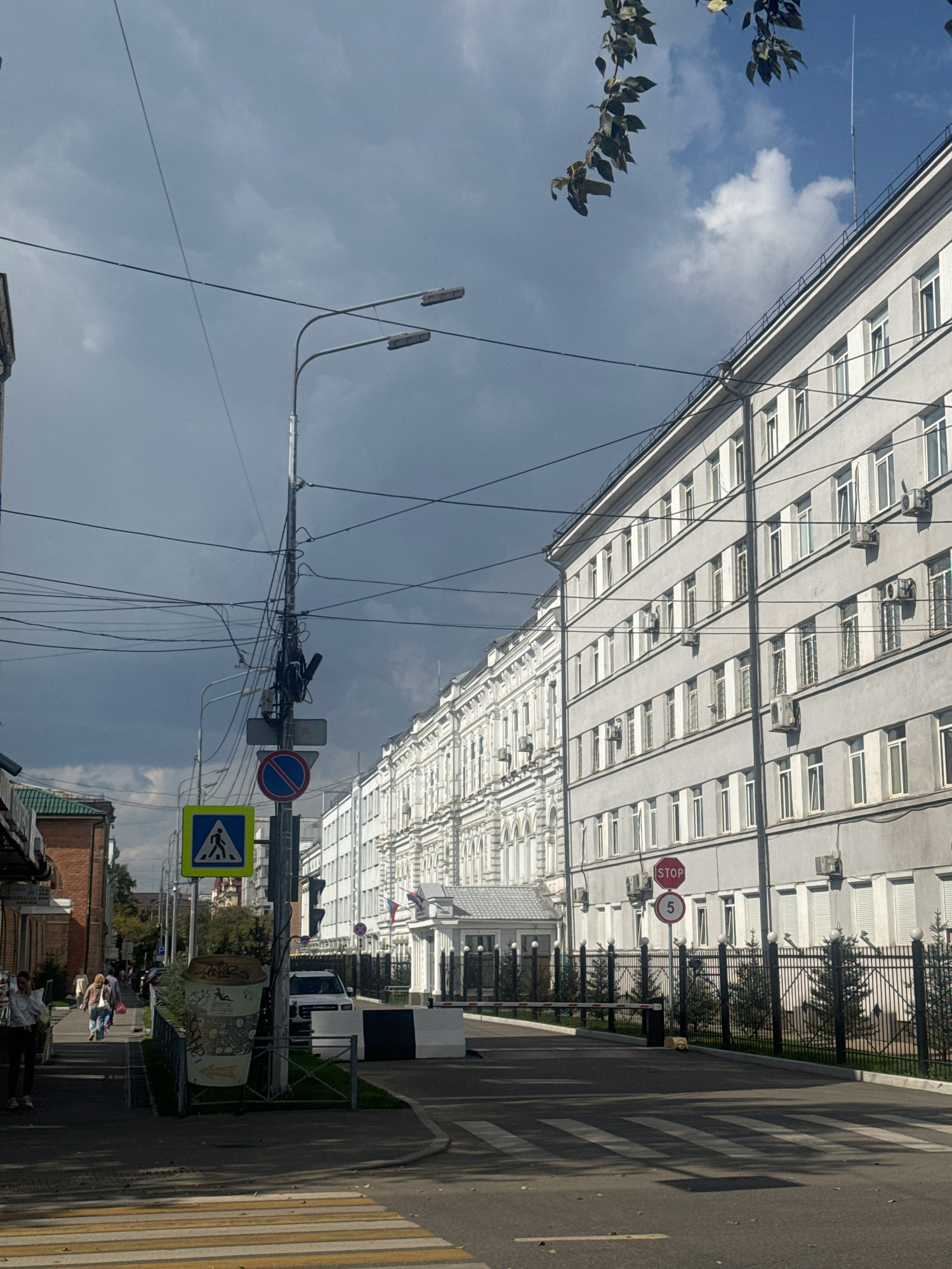 The image depicts a street scene in the city of Irkutsk, Russia, likely in an urban or semi-urban area. The perspective is from street level, looking towards a large, modern, multi-story building with a light-colored facade and numerous windows. The building appears to be an institutional or commercial establishment, possibly an office building or administrative facility. In front of this building, a black metal fence with decorative elements encloses the property, and a road barrier is visible, suggesting controlled access. Traffic signs are present, including a "STOP" sign and a sign with the number "5" indicating a speed limit.
To the left of the main building, a pedestrian crossing is marked on the asphalt road. A pedestrian signal and a "Pedestrian Crossing" sign are visible on a utility pole heavily laden with overhead wires. Further down the street on the left, there are other buildings, one with a dark brick facade and a green roof, and more people walking along the sidewalk. The sky is partly cloudy, with patches of blue sky visible amidst darker, heavier clouds.
Several individuals are present in the scene, mostly walking along the sidewalk on the left side of the image. One person is in the foreground on the left, walking away from the camera. A few other figures are visible further down the sidewalk. The overall impression is that of a typical day in a Russian city, with a mix of modern and older architecture, infrastructure, and pedestrian activity. The presence of a large building with a barrier and security features might indicate a government office or a significant business.