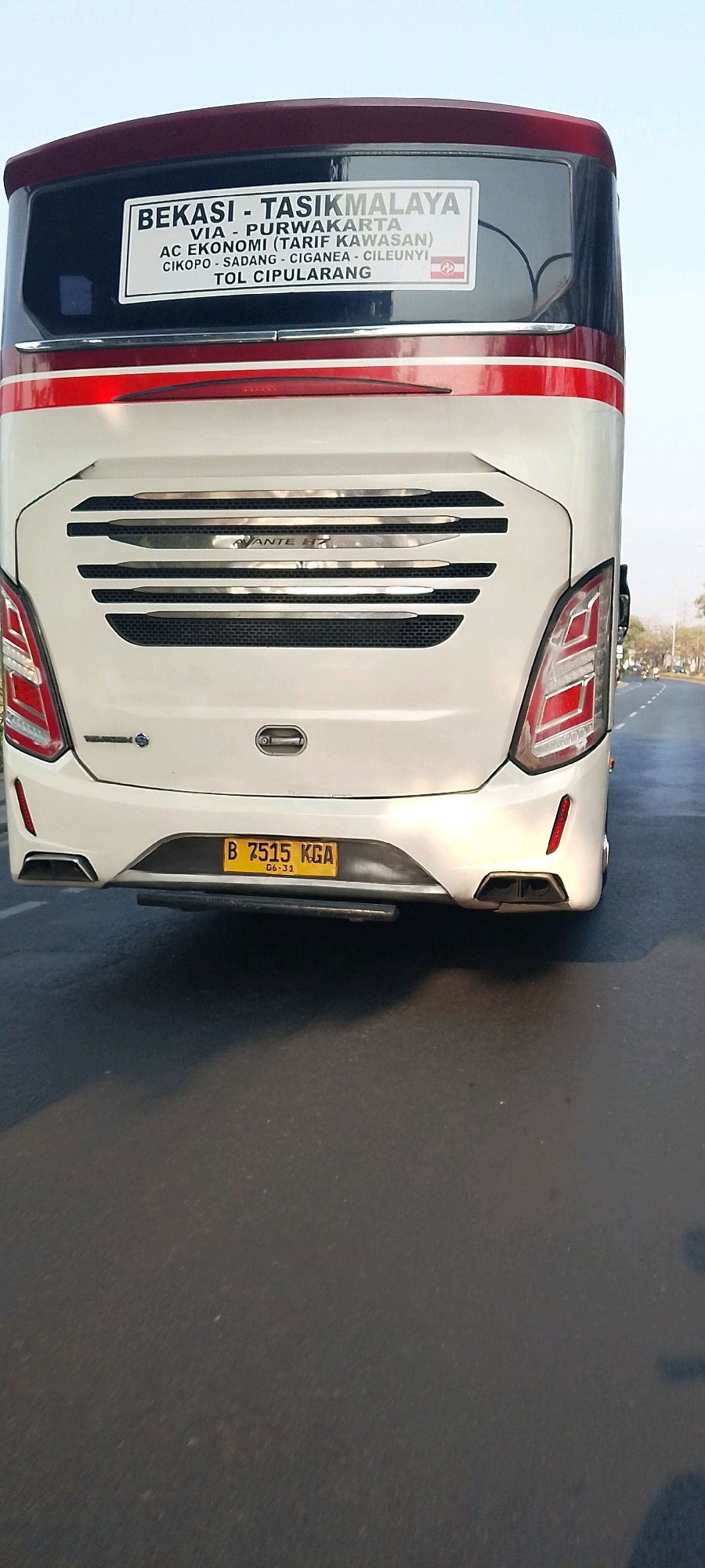 The image shows the rear of a white bus with red and black accents. A sign on the back of the bus reads "BEKASI - TASIKMALAYA VIA - PURWAKARTA AC EKONOMI (TARIF KAWASAN) CIKOPO - SADANG - CIGANEAD - CILEUNYI TOL CIPULARANG". The license plate reads "B 7515 KGA". The bus appears to be driving on a road, possibly in Bekasi, Indonesia, given the sign. The environment is outdoors with a clear sky.