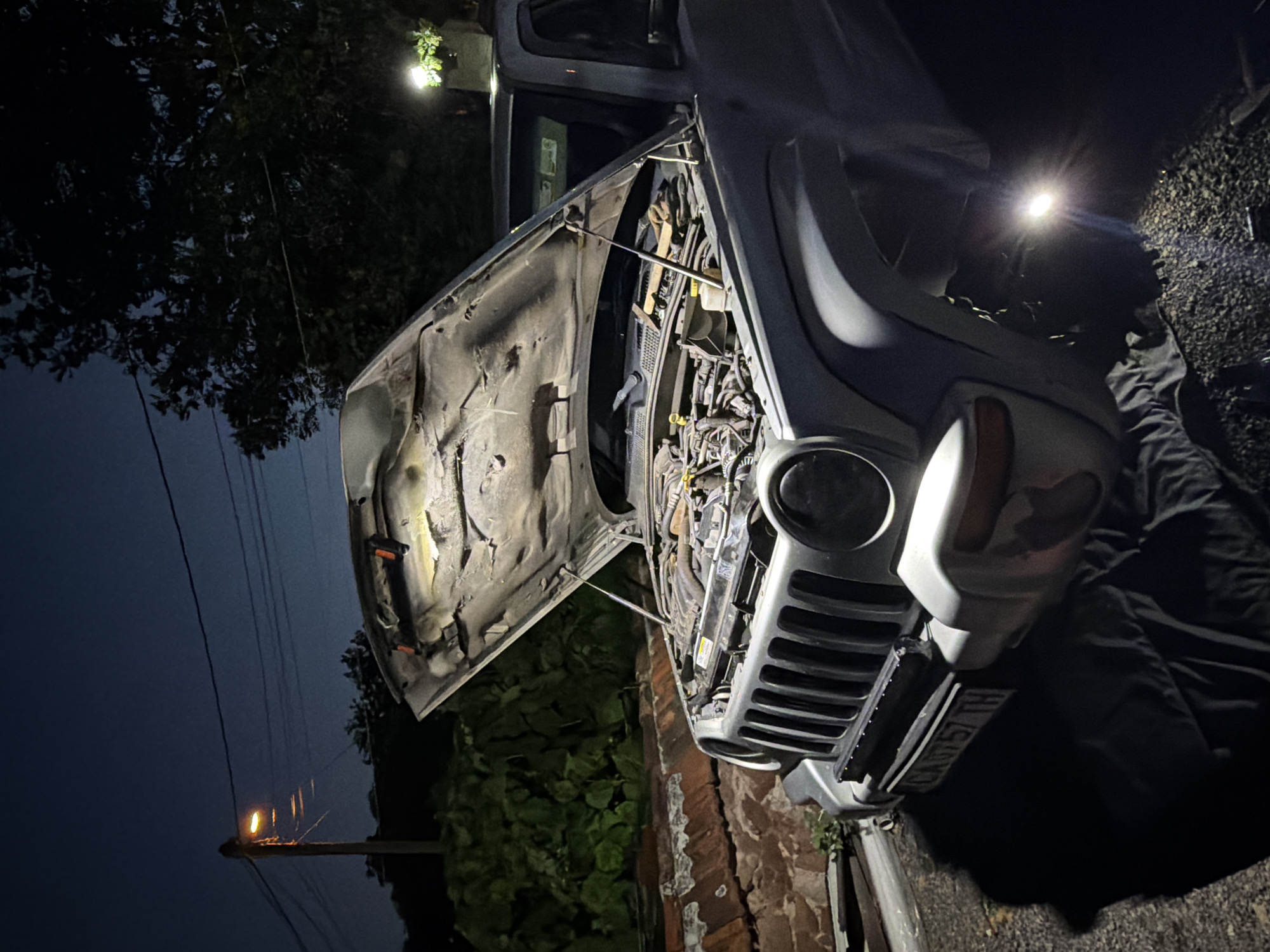 In the city of Voynegovtsi, Bulgaria, at night, a silver Jeep Liberty is parked with its hood open, revealing the engine. The vehicle is positioned on a dark, gravelly surface, with a black tarp spread underneath it. A brick wall and dense green foliage are visible behind the car. A street lamp with a warm, orange glow stands to the left, illuminating power lines that stretch across the dark blue sky. Additional artificial lighting, possibly from a work lamp placed on top of the Jeep, highlights the engine compartment. The license plate of the Jeep reads "CA 0757 TH". The overall scene suggests a roadside repair or maintenance operation taking place after dark.