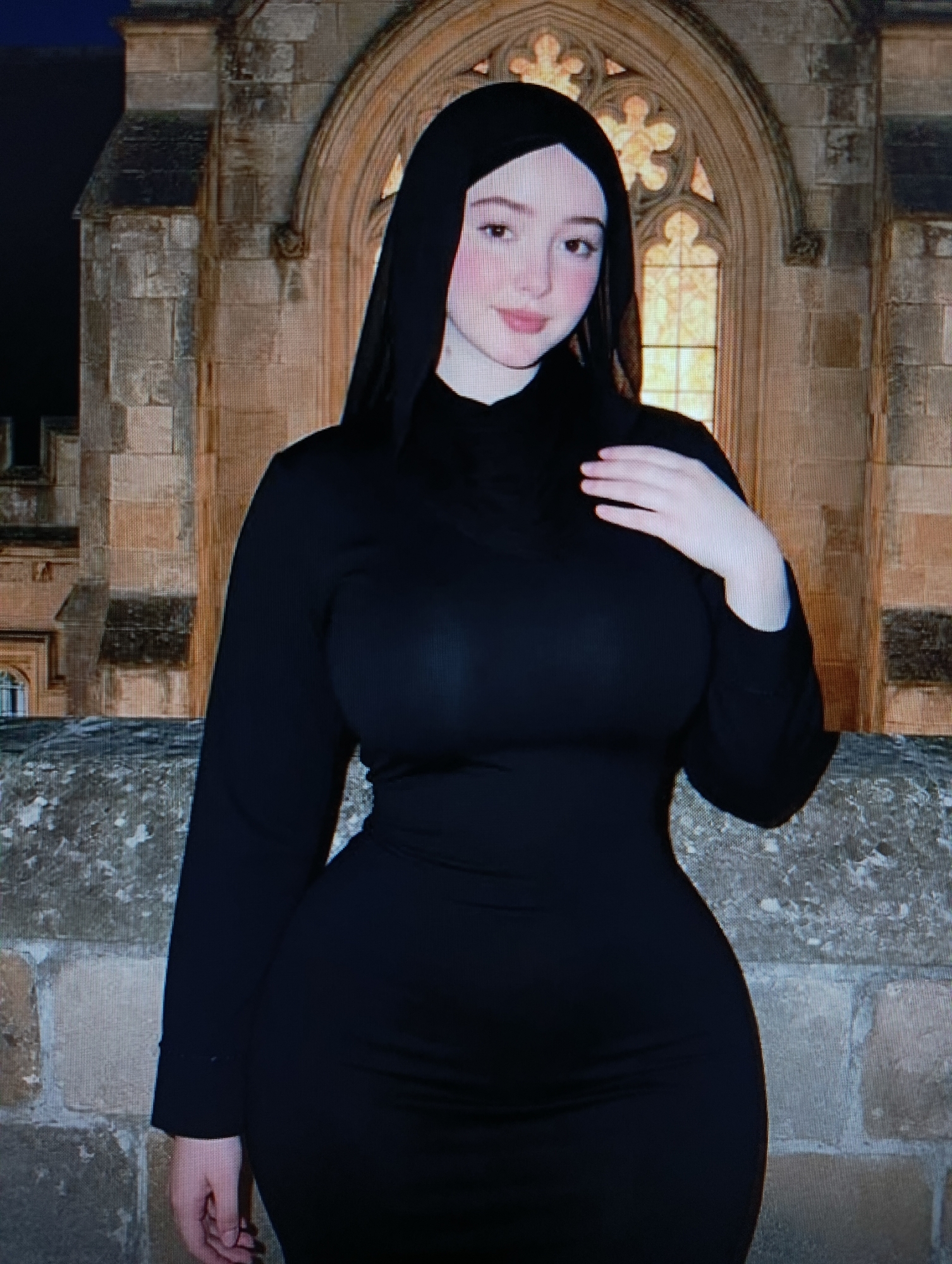 A young woman, wearing a black hijab and a long-sleeved black dress, stands in front of an old stone building with a Gothic-style window. Her left hand is placed on her chest, and her right hand rests by her side. The background features weathered stone walls and a dimly lit sky.