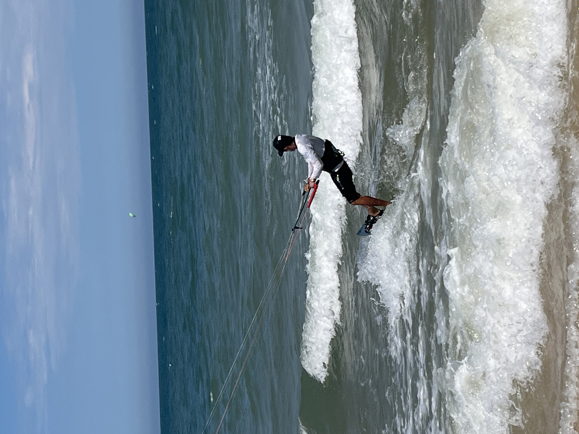 In the ocean off the coast of Phu Thuy Ward, Vietnam, a person kiteboards on a sunny day. The individual, wearing a white long-sleeved shirt, black shorts, a black cap, and a kiteboarding harness, is actively engaged in the sport. They are holding onto a control bar attached to kite lines that extend upwards and out of frame. The kiteboarder is positioned on a board in the water, with waves breaking around them. In the distance, another smaller kite is visible in the sky, along with a few distant figures in the water. The ocean is a deep blue-green, with white foam from the waves in the foreground. The sky is a pale blue with some scattered clouds.