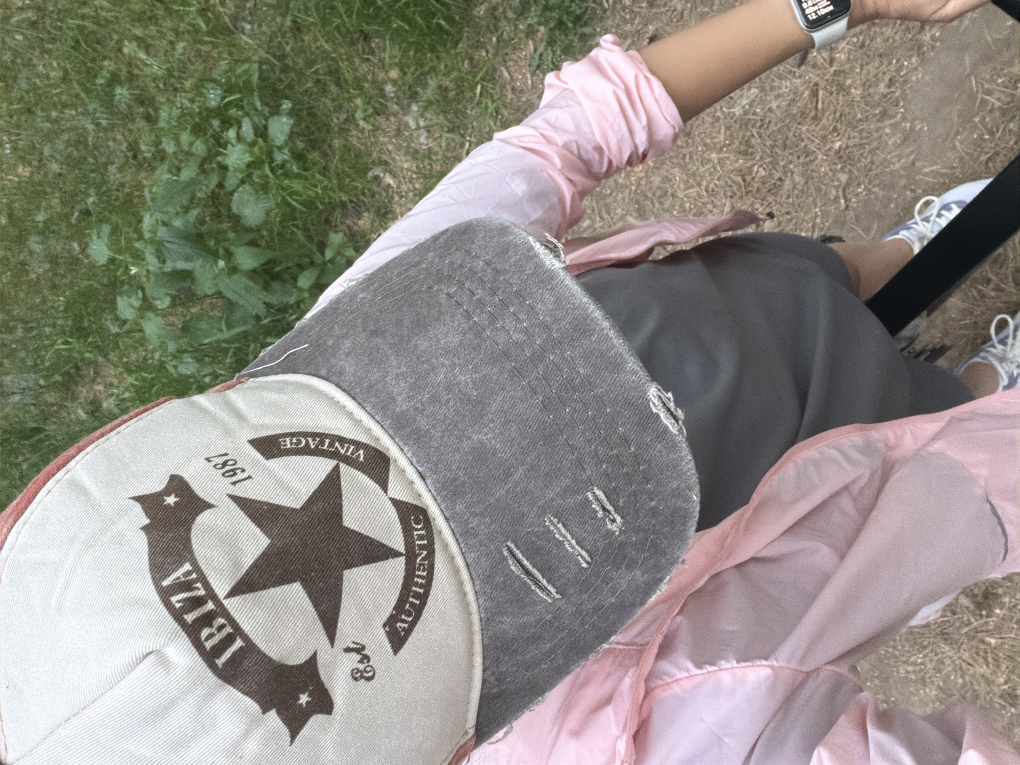The image is a top-down, close-up view. The person is wearing a light pink jacket and a gray and beige baseball cap with "IBIZA," "Vintage 1987," and a star design. The person is also wearing an Apple Watch on their left wrist. The person is wearing a gray skirt, pink jacket, and blue tennis shoes with white laces. The background includes grass and dry leaves/straw. The location cannot be verified.