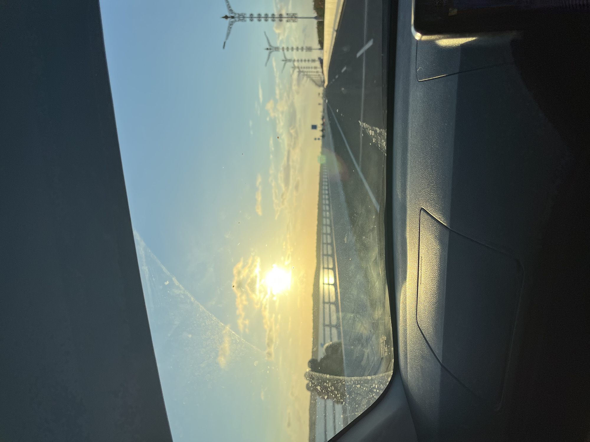 The image shows a view from inside a vehicle, likely a car, driving on a road in Kuta Selatan, Indonesia. The sun is low in the sky, appearing to be setting or rising, and shines brightly through the windshield. The road is straight and appears to be a highway or major roadway. In the distance, there are tall, decorative structures, possibly light poles or towers. The sky is blue with some scattered clouds. The dashboard of the car is visible, occupying the right side of the frame. There is some dirt or smudges on the windshield.