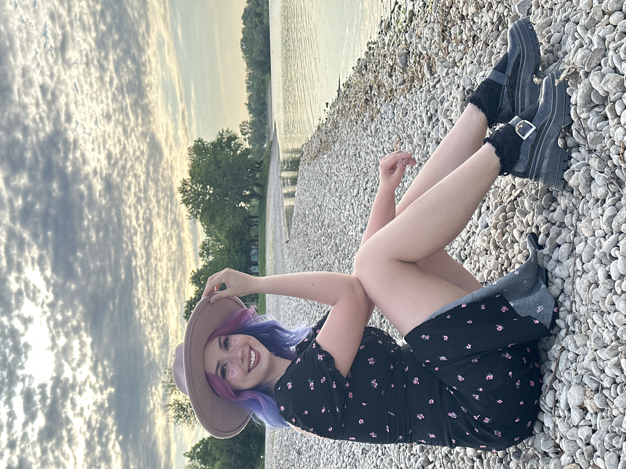A young woman is depicted lying on her side on a pebbled shore, smiling and posing for a photograph. She is wearing a black dress adorned with small pink floral patterns, dark ankle boots with buckles, and black frilly socks. Her long, wavy hair is dyed in shades of purple and blue. She has small heart-shaped glitter details under her eyes and is wearing a light brown wide-brimmed hat, which she holds with one hand.
The setting is a waterfront location, with a body of calm water stretching out behind her, reflecting the sky. On the opposite side of the water, a line of trees and greenery is visible, suggesting a natural landscape. The shore consists of numerous light-colored pebbles and small stones.
The sky is filled with a pattern of white and grey clouds, possibly altocumulus or stratocumulus, with areas of brighter light breaking through, indicating either sunrise or sunset, or soft afternoon light. The overall mood is relaxed and joyful, conveyed by the woman's smiling expression. There is no visible text in the image. The scene likely takes place at a lake or river within or near the City of Zagreb, Croatia.