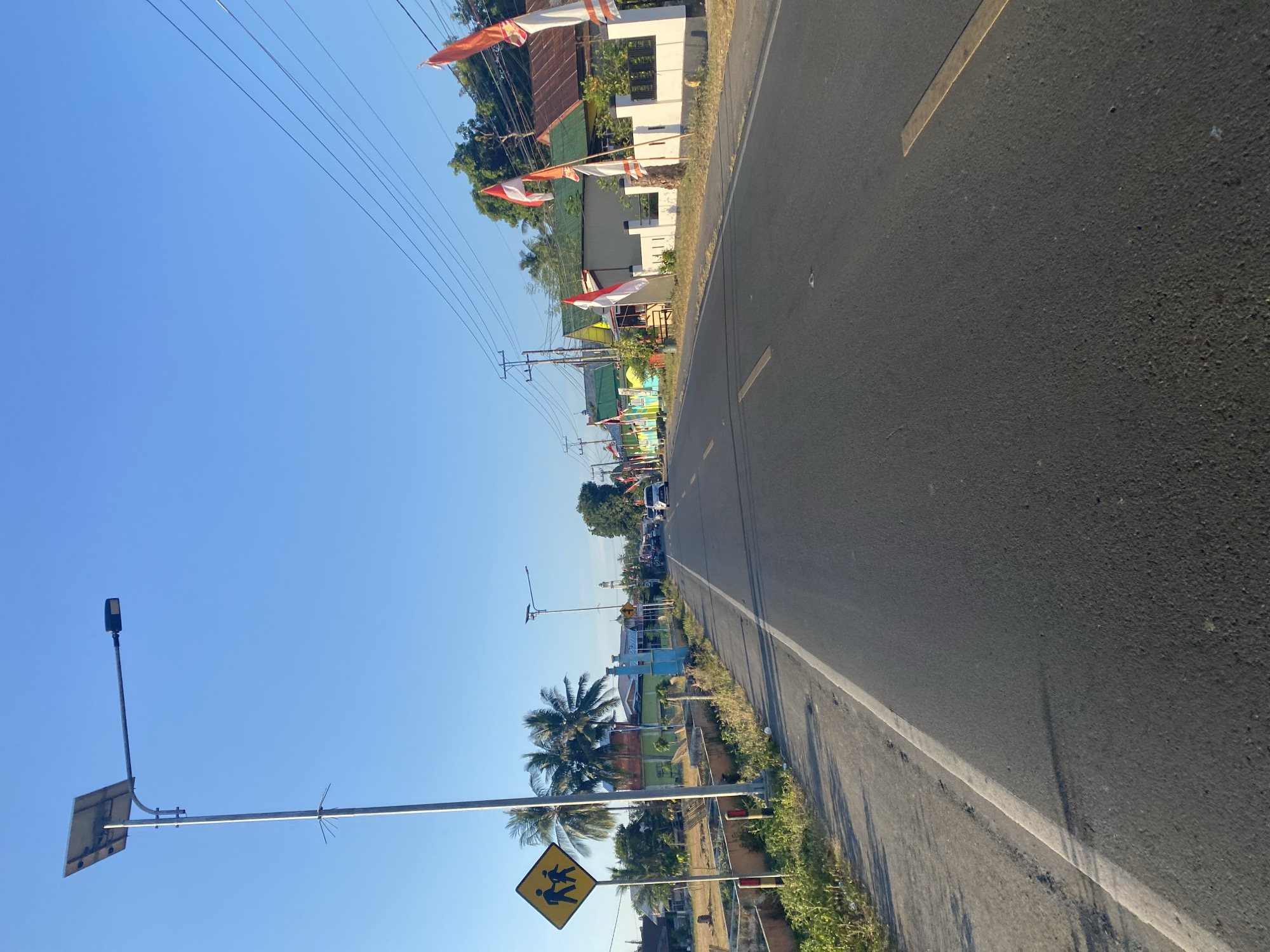 The image depicts a street scene in Palambarae, Indonesia. A two-lane asphalt road runs through the frame, with the perspective suggesting the viewer is moving along the road. On the left side of the road, a yellow diamond-shaped pedestrian crossing sign with two silhouetted figures is visible, mounted on a tall pole that also supports a solar-powered street light. Adjacent to the sign, palm trees and some vegetation can be seen.
Along the right side of the road, a row of buildings with colorful facades and corrugated metal roofs are present. Many of these buildings are adorned with Indonesian flags, indicating a possible celebration or national holiday. Power lines stretch across the sky above these structures. Further down the road, several vehicles, including cars, are visible. The sky is clear and blue, suggesting a sunny day. The overall environment appears to be a semi-rural or suburban area with residential buildings and infrastructure.