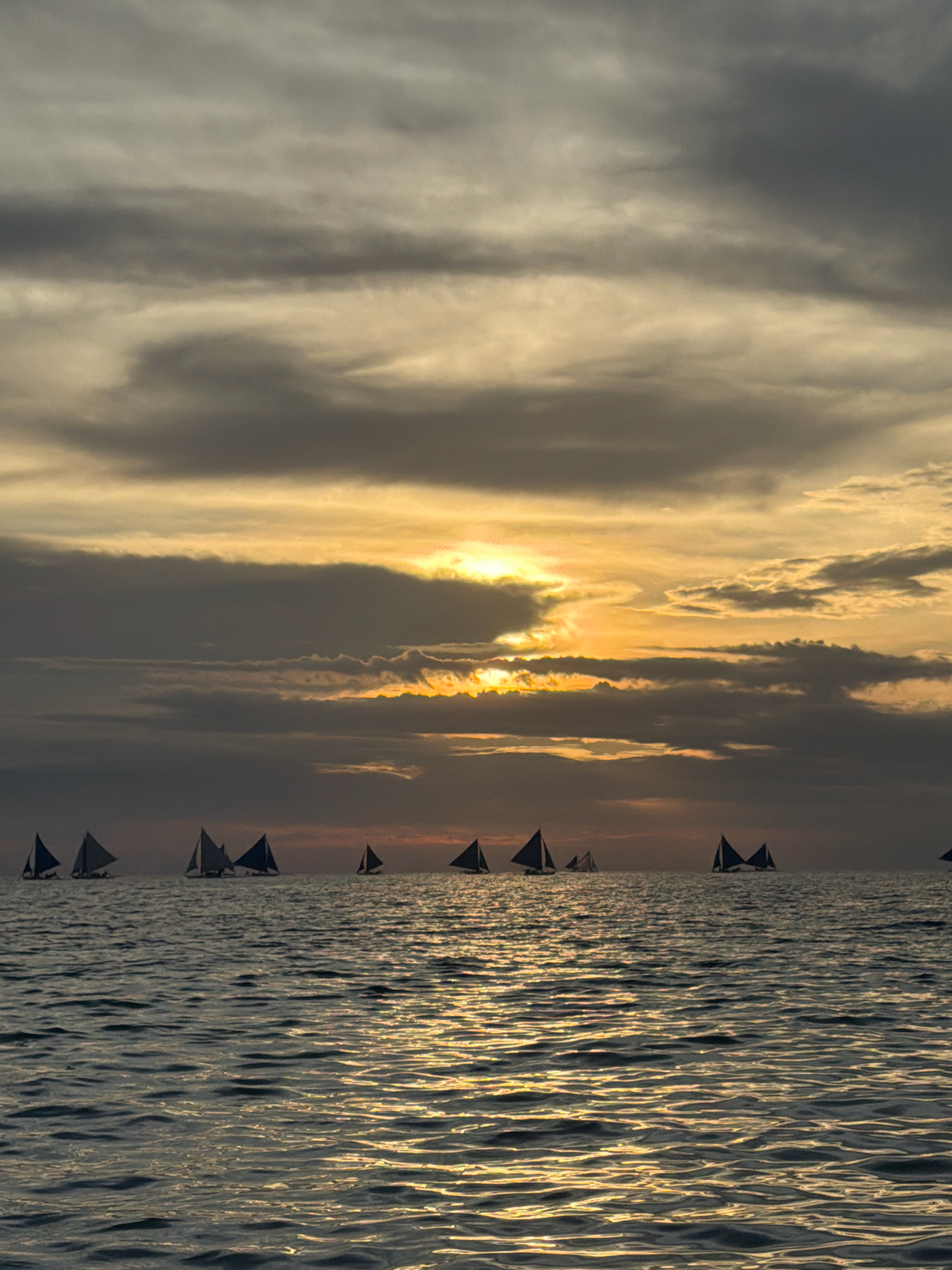 The image captures a sunset over the sea in Malay, Philippines, likely Boracay. A fleet of sailboats with triangular sails dots the horizon. The sky is filled with dramatic, layered clouds, with the setting sun peeking through, casting a warm, golden glow on the water. The ocean surface reflects the colors of the sky, creating a shimmering effect. The scene evokes a sense of tranquility and natural beauty.