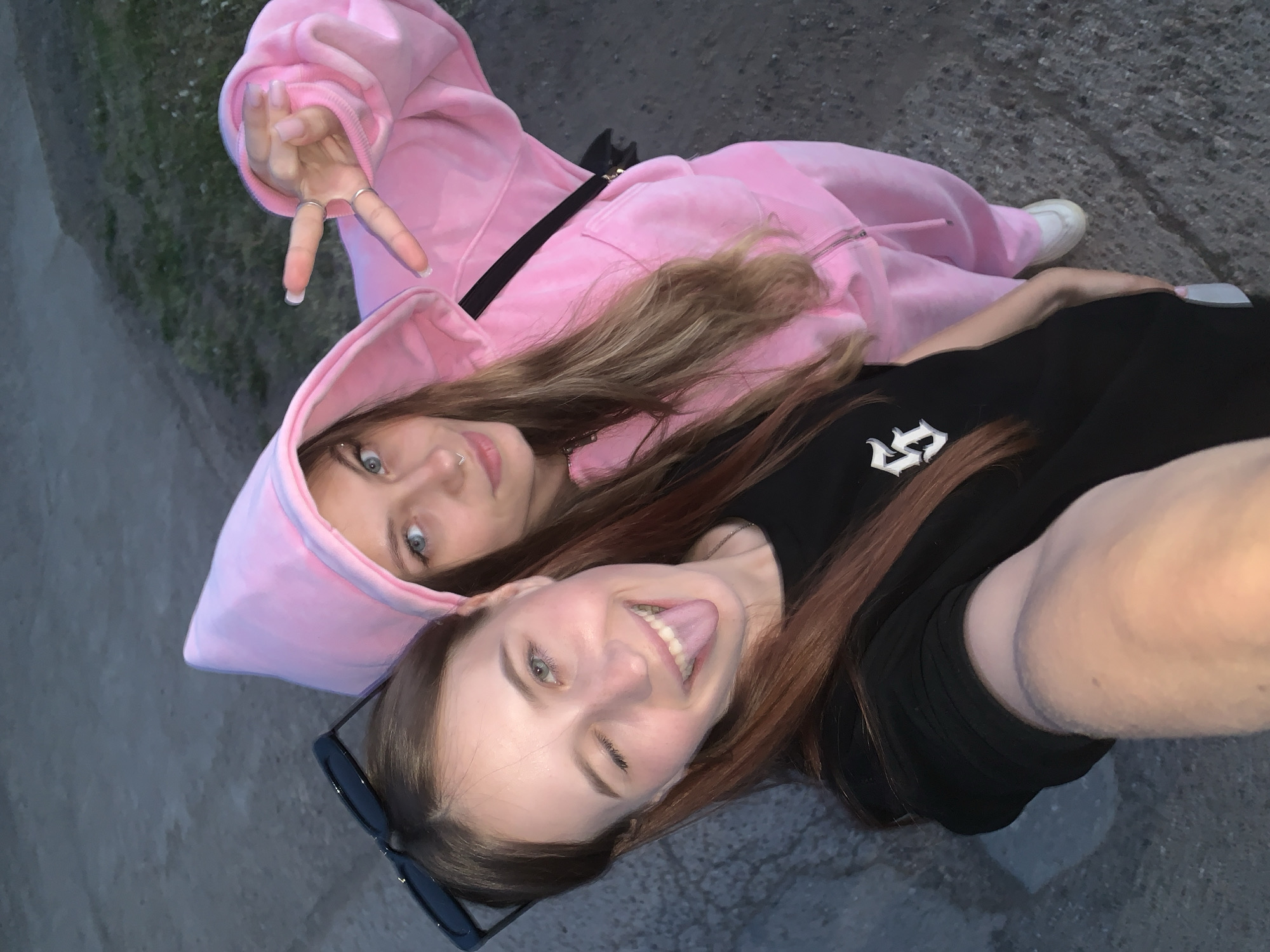 Two young women are lying on a concrete surface, likely a sidewalk or paved area. The woman on the left wears a light pink hooded sweatsuit and makes a peace sign with her hand. The woman on the right wears a black t-shirt with a white logo, sticks out her tongue, and winks. A pair of sunglasses rests beside her. The background appears to be urban. It is not possible to confirm the location is in Фоки, Russia from the visual information provided.