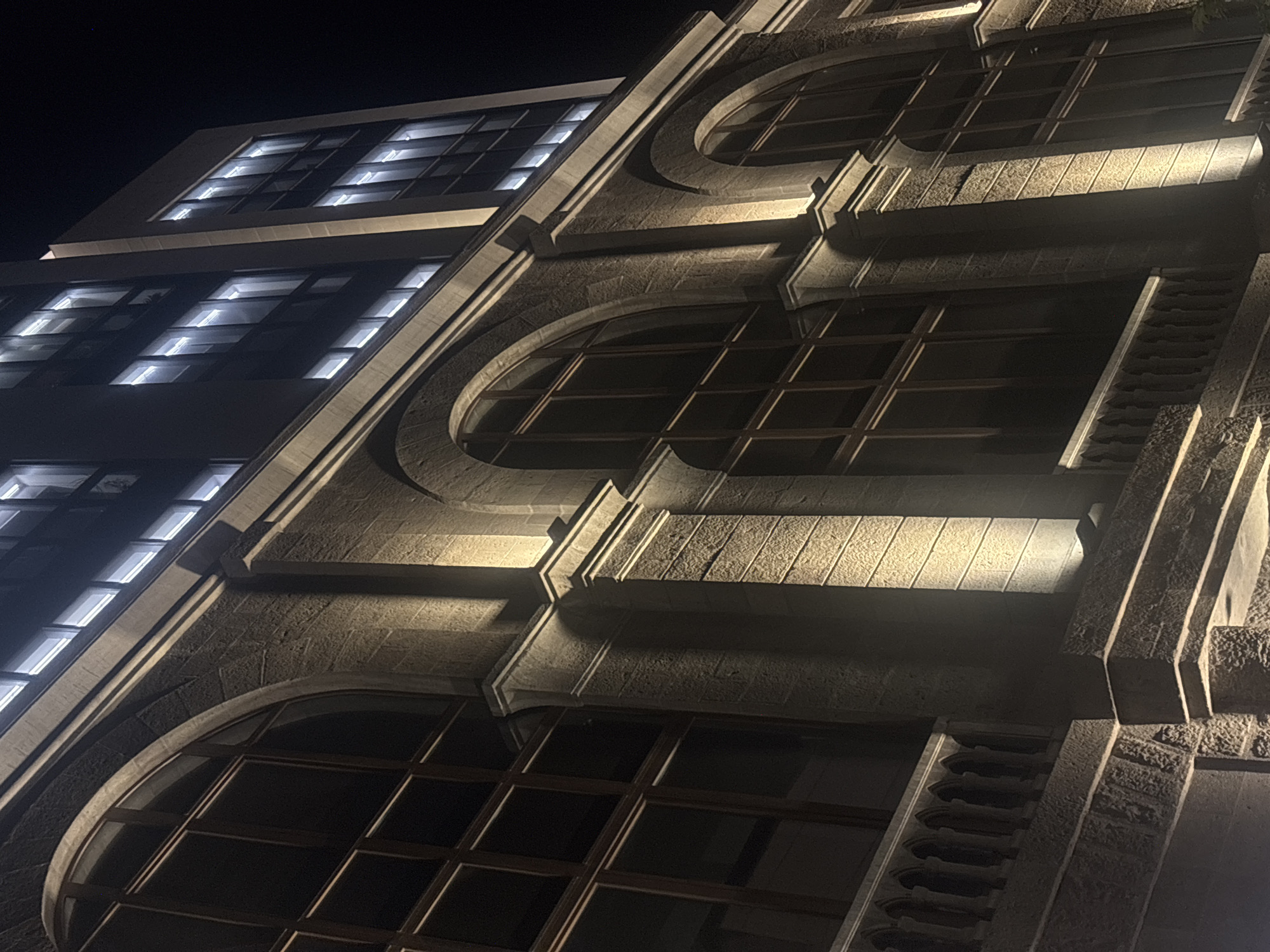 The image shows a building facade, likely in Yerevan, Armenia, given the architectural style. The building's exterior is stone, illuminated by artificial light, which casts shadows and highlights the texture of the stone. There are windows on the left side, which are rectangular, and windows on the right side, which are oval-shaped with grid patterns. The stone facade appears to have decorative elements and detailing, with multiple levels and recesses. The sky in the background is dark, suggesting it is night time.