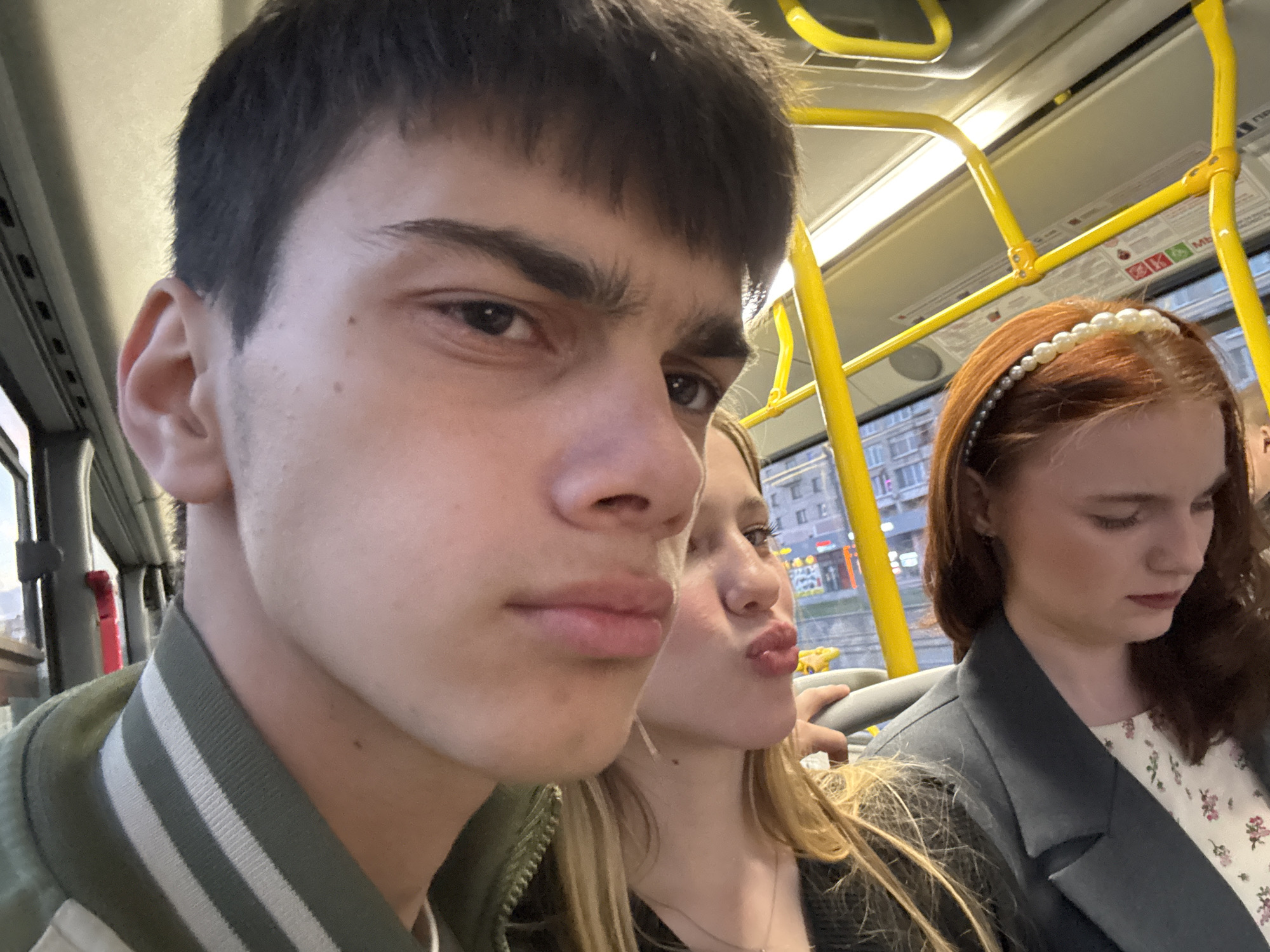 A selfie-style photo captures three young people inside what appears to be a public transport vehicle, likely a bus or trolleybus, in an urban setting during dusk or evening.
In the foreground, a young man with dark, short hair and dark eyebrows looks directly at the camera with a neutral expression. He is wearing a dark green jacket with white stripes on the collar. Behind him, to his right, is a young woman with long blonde hair, puckering her lips in a playful expression. Behind her, and slightly to the right, is another young woman with auburn hair, adorned with a white pearl headband. She is looking downwards or to her right, appearing contemplative, and is wearing a dark jacket over a white top with a floral pattern.
The interior of the vehicle features bright yellow grab handles and poles, typical of public transport in many cities. Through the windows, blurred buildings with some illuminated windows and streetlights are visible, indicating an urban environment at twilight.
Above the head of the woman with the pearl headband, a sign on the vehicle's interior features some partially visible Cyrillic text, including "МБ" and what appears to be "При", along with some small red and green icons. This, combined with the location context, suggests the scene takes place in Russia. The overall activity is a candid moment of friends traveling together and taking a picture.