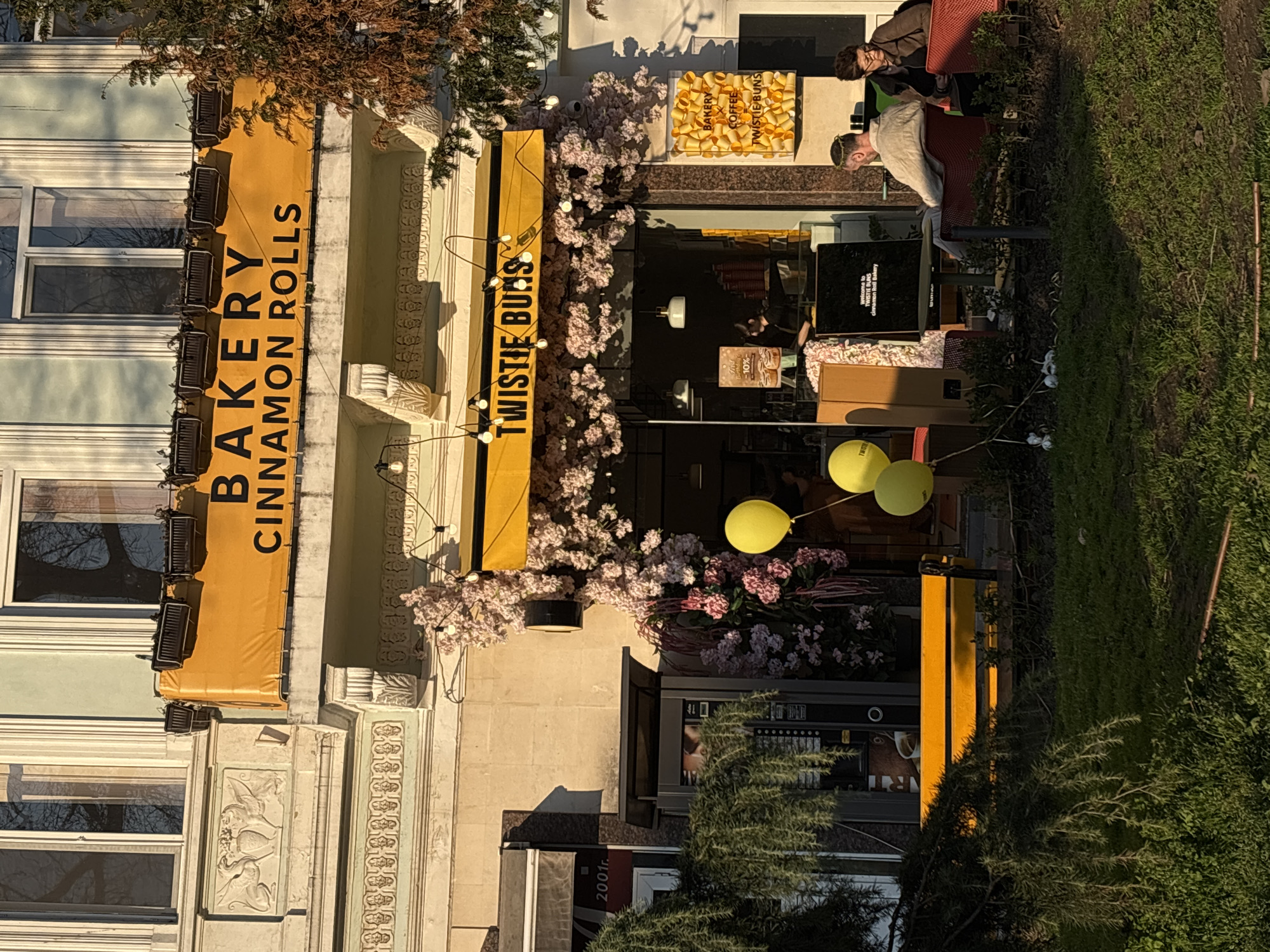 The image shows the exterior of "Twistie Buns," a bakery in Varna, Bulgaria. The building has a yellow banner with "Bakery Cinnamon Rolls" written on it, and another sign saying "Twistie Buns." Pink flowers decorate the storefront. A display of Twistie Buns is visible in the window. Outside, there are yellow balloons and seating where people are sitting and standing. There is greenery alongside the building.