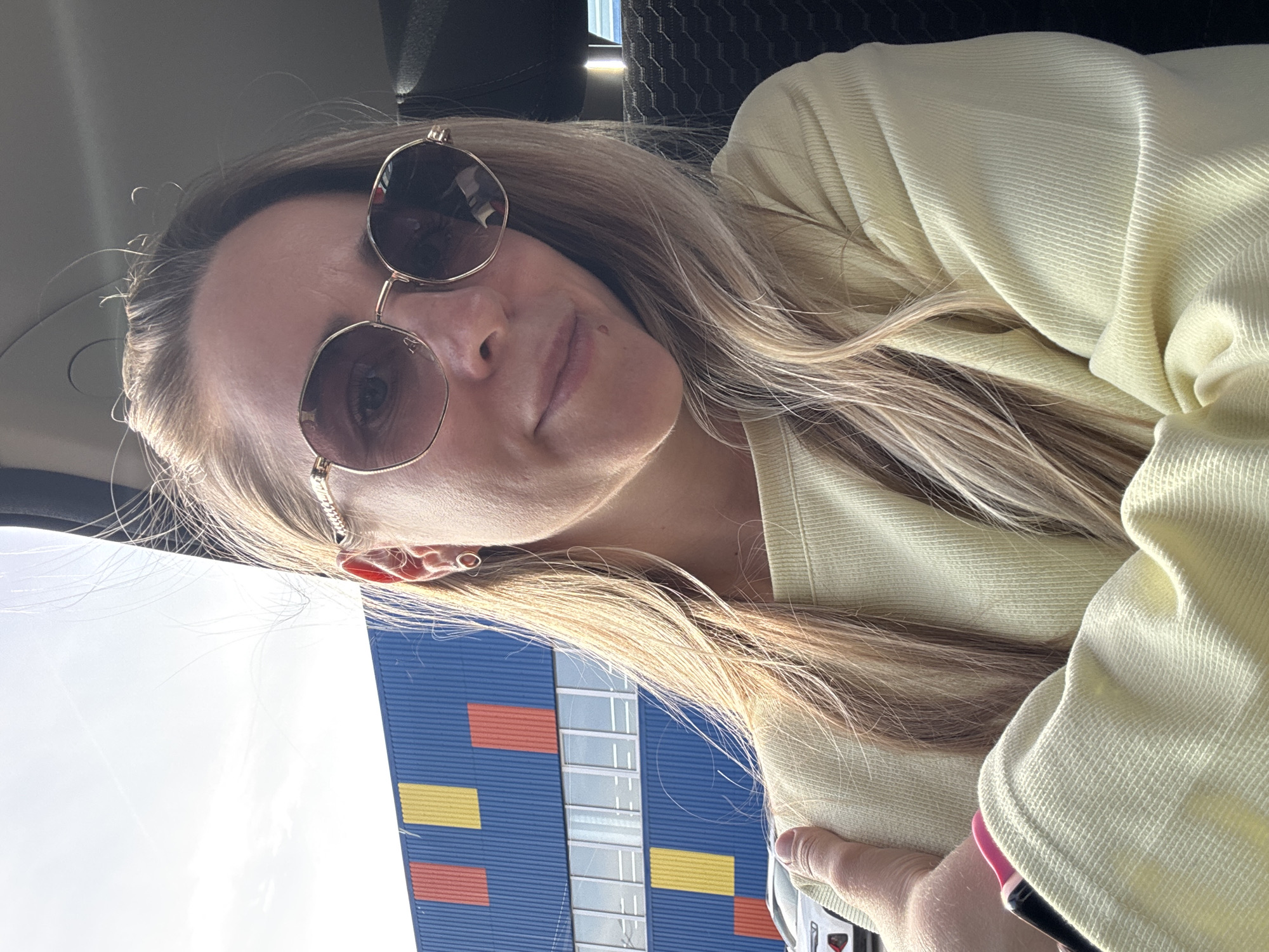 A woman with blonde hair and round sunglasses is photographed from a low angle. She is wearing a light yellow shirt or sweater. Behind her, a tall building with a blue facade and orange and yellow rectangular accents is visible. The architecture style and the building's colors indicate that it is likely an IKEA store. Given the prompt's location, this suggests the photo may have been taken in or near the IKEA store within the city of Chaikovsky, Russia. The angle suggests she's possibly sitting in a car, given the car interior visible in the background.