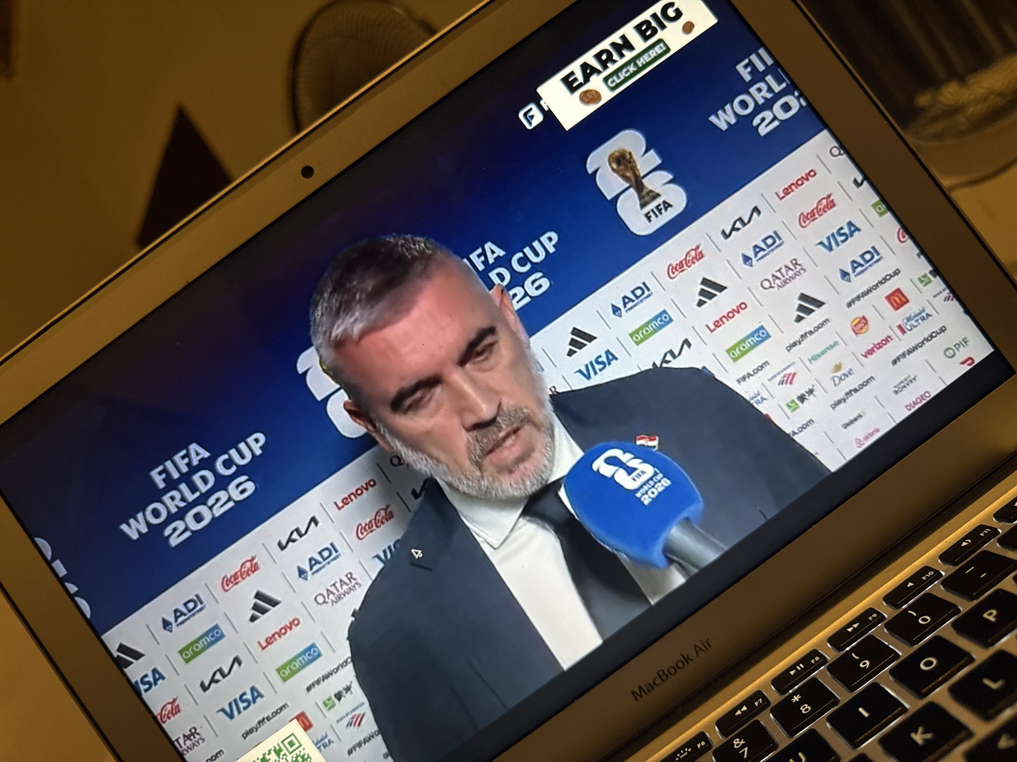 The image shows a close-up, angled view of a MacBook Air laptop screen displaying a broadcast related to the FIFA World Cup 2026. The screen shows a man in a dark suit and tie, speaking into a blue microphone with the FIFA World Cup 2026 logo on it. Behind him is a digital backdrop with numerous sponsor logos, including Adidas, Visa, Coca-Cola, Qatar Airways, and Lenovo. A promotional banner on the right side of the screen reads "EARN BIG" with the text "CLICK HERE!". The laptop keyboard is partially visible in the foreground. The environment appears to be indoors, with warm lighting suggesting evening or nighttime. There is no indication of the city of Kufena, Nigeria in the image.